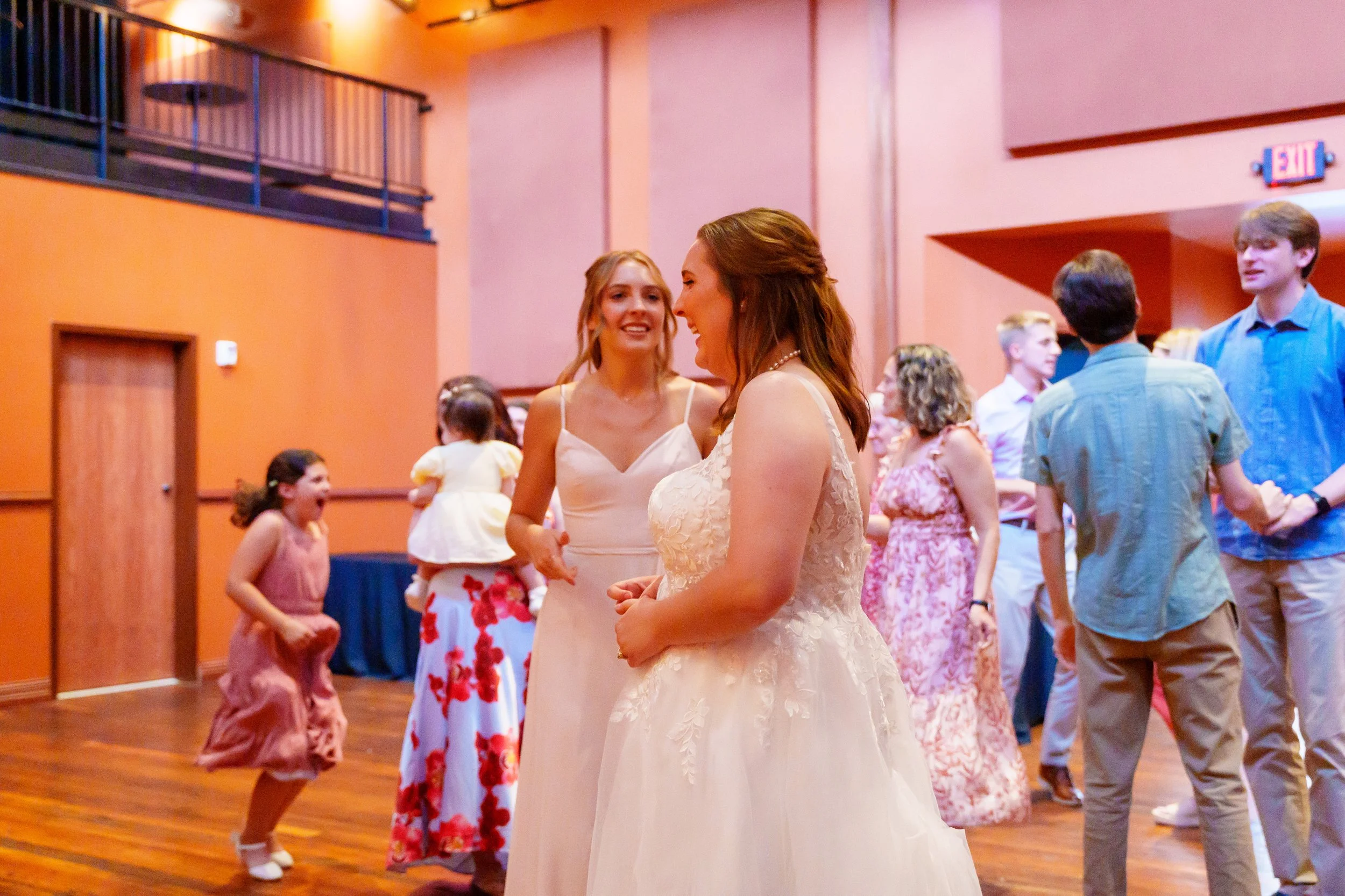 People dancing at a social event, with two women in the foreground talking, one in a white dress and the other in a pink dress. The background shows other attendees, including children, and a room with pink walls, a wooden floor, and an exit door.
