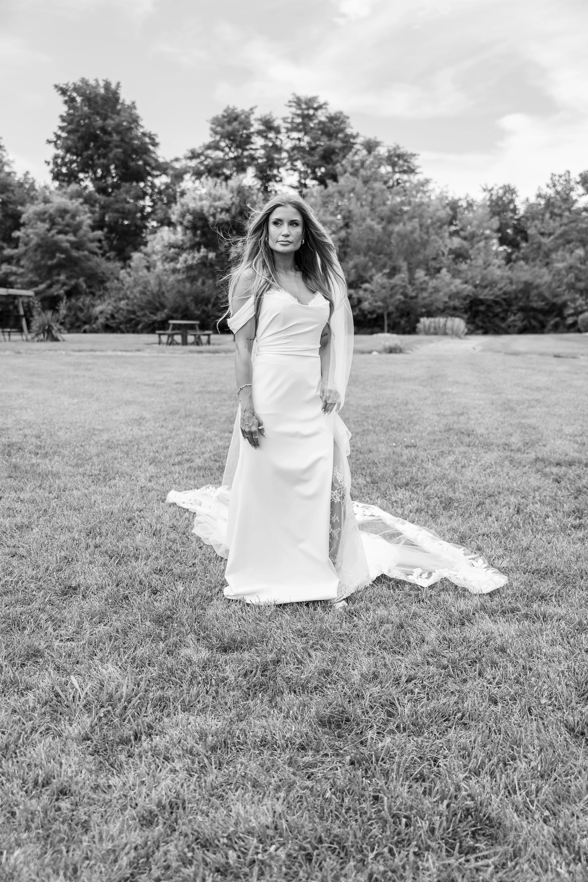 A woman in a wedding dress standing on a grassy field with trees in the background, looking off to the side.