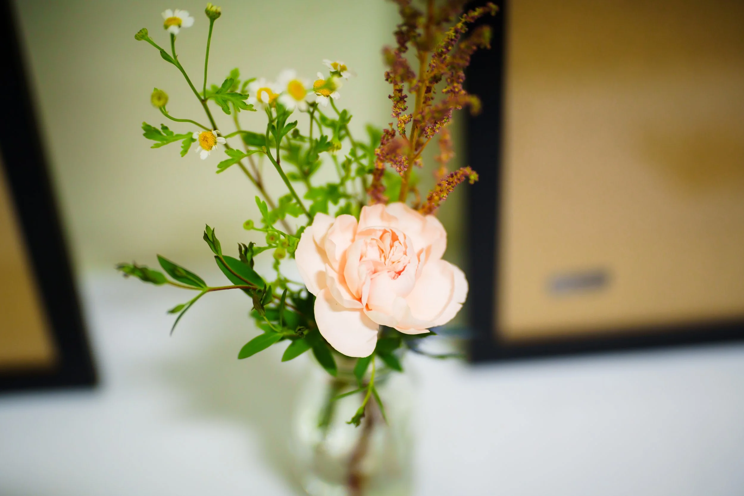 A close-up of a floral arrangement with small white and yellow daisies, a pale pink rose, and purple-tinted greenery on a white surface.