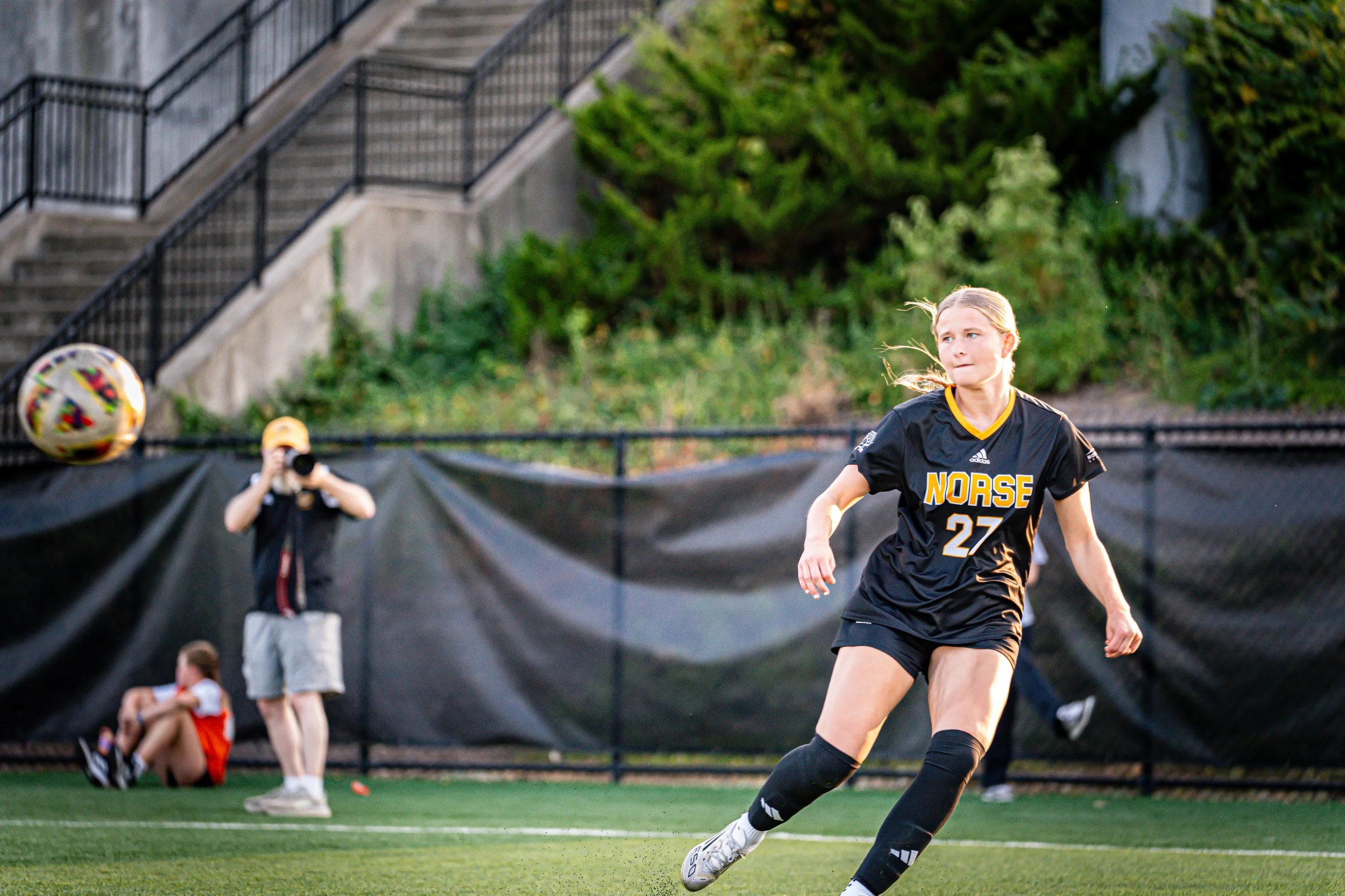 A girl in a black soccer uniform with the number 27 and the word 'Norse' on her jersey is running on a soccer field, with her hair flowing behind her. In the background, a person is taking photos or videos with a camera, and a young girl in a red and
