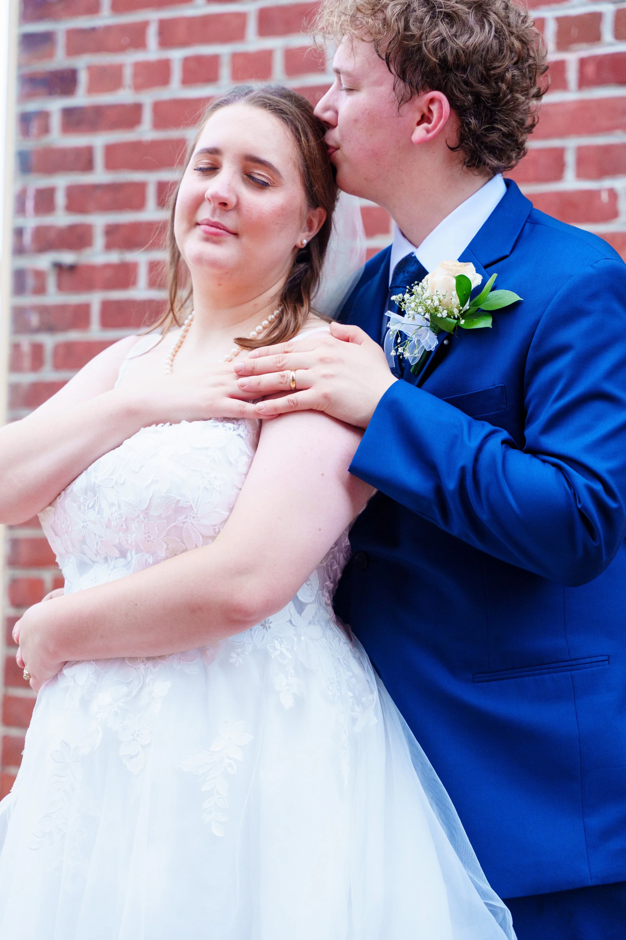 A bride and groom embrace in front of a red brick wall. The groom is kissing the bride on the side of her head. The bride is wearing a white wedding dress with lace details and a pearl necklace, and the groom is in a bright blue suit with a white shirt, dark tie, and a boutonniere. The bride's eyes are closed, and she looks content.