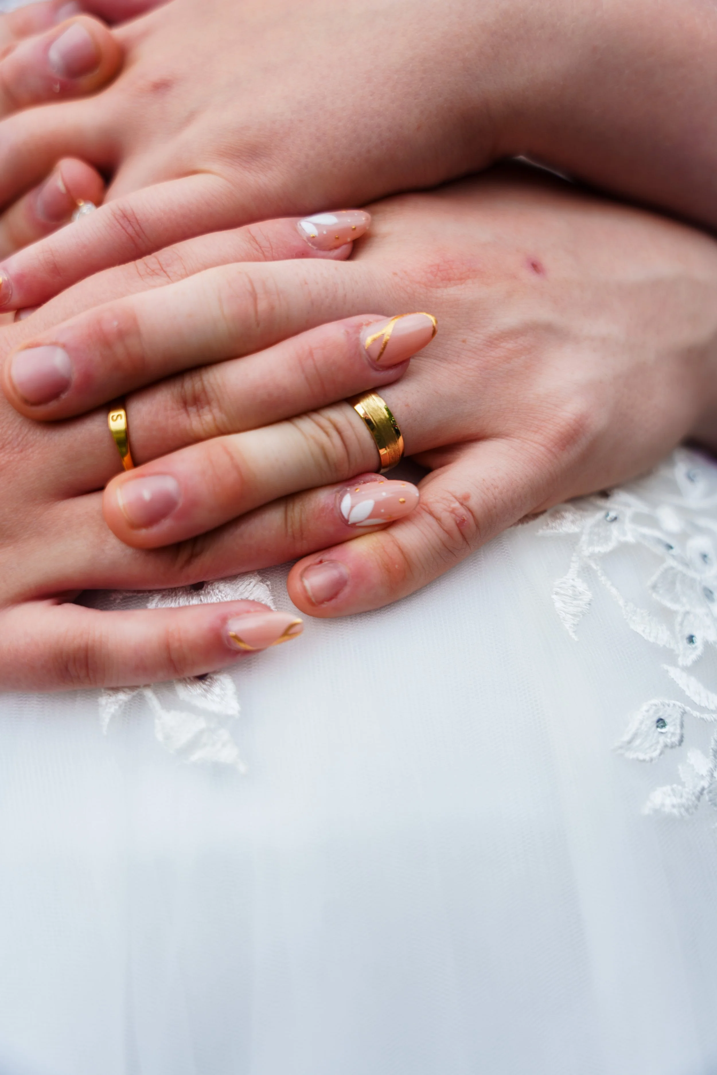 Close-up of two hands, one with a gold wedding ring, resting on a white fabric with embroidered details.
