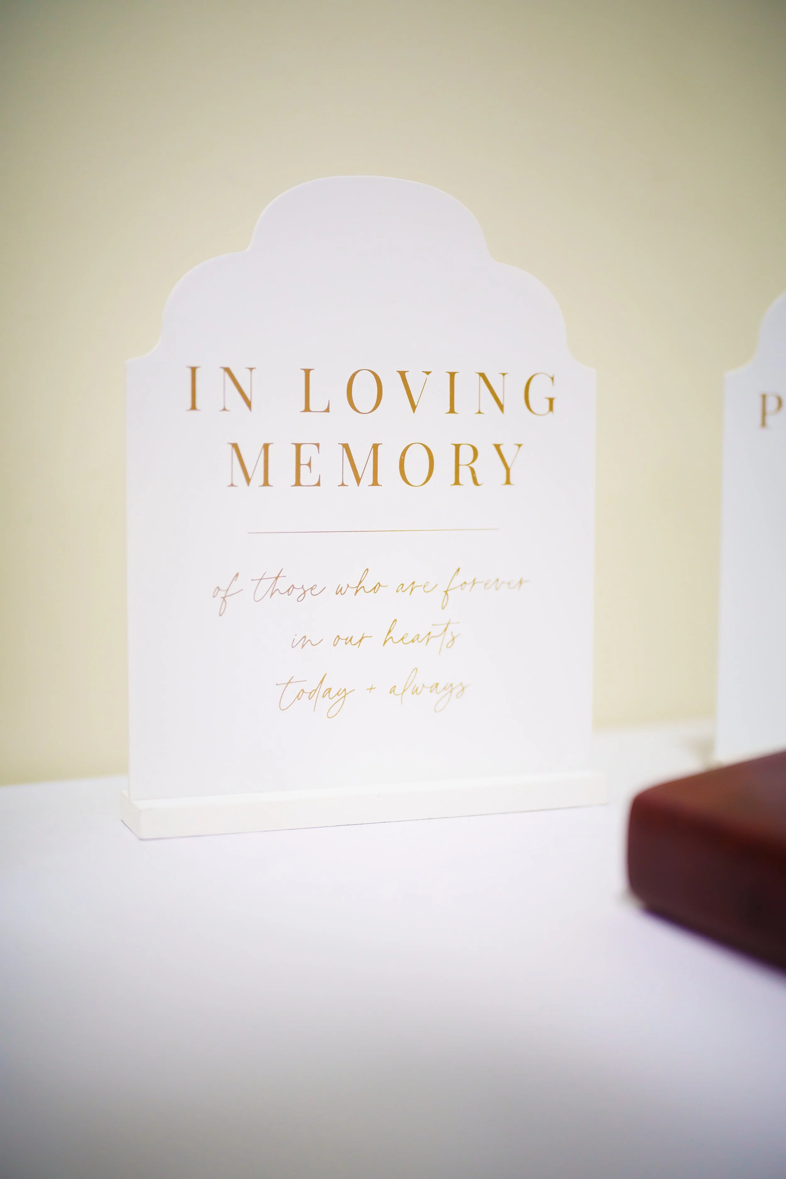 A white decorative sign with gold lettering that reads 'In Loving Memory' and additional cursive writing underneath, positioned on a white surface with a blurred background.