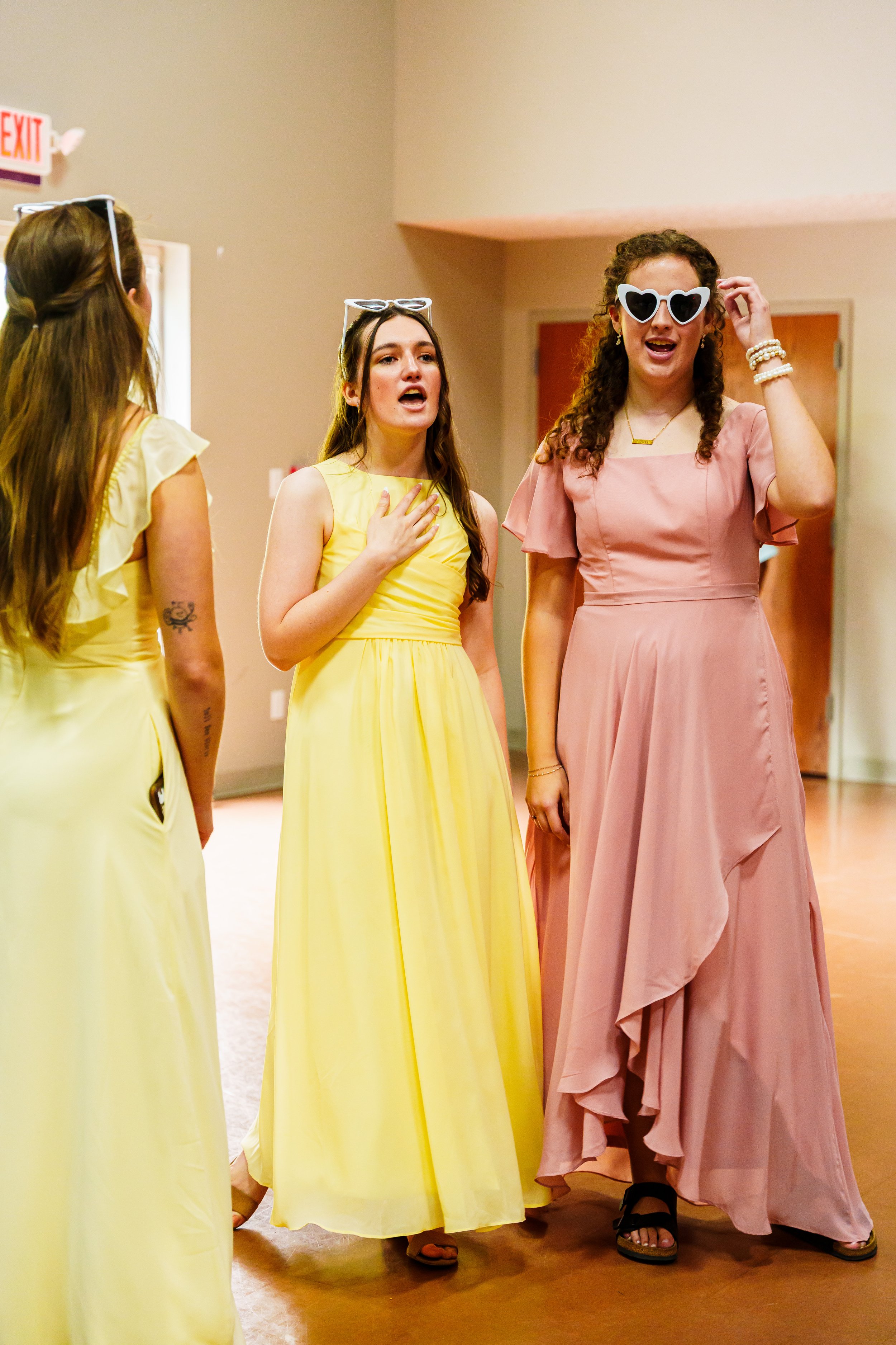 Four women dressed in pastel-colored evening gowns engaged in conversation at a social event in an indoor hall.