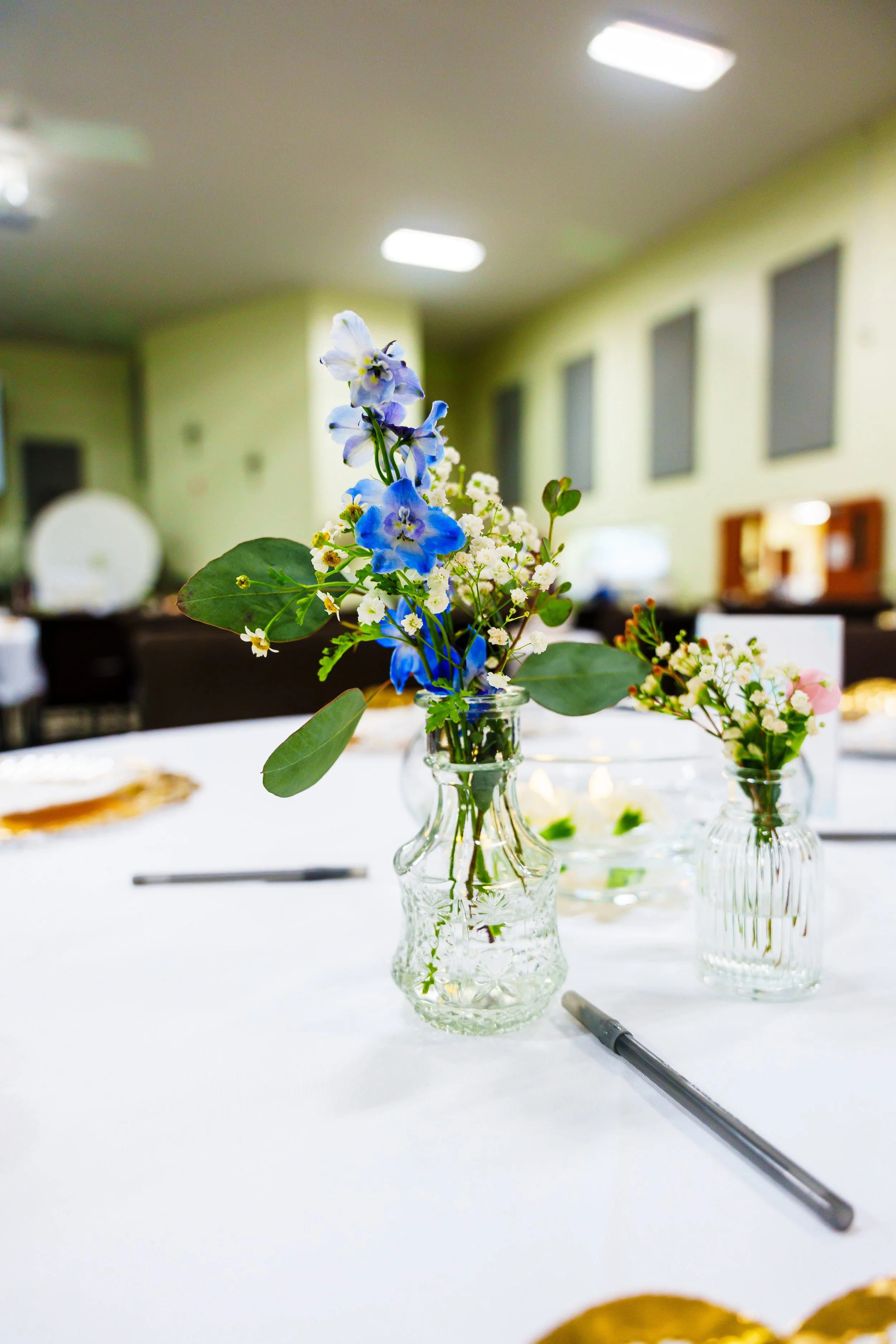Close-up of three small glass vases with blue and white flowers on a white table in a room with fluorescent ceiling lights and windows.