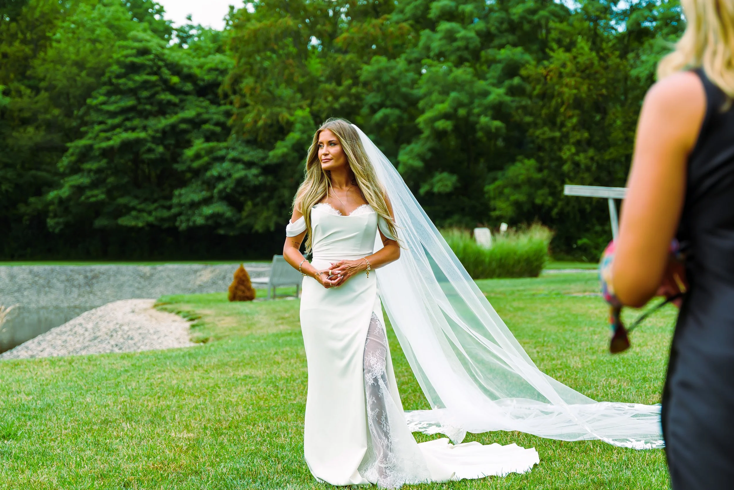 A bride in a white wedding dress with lace detailing and a long veil, standing outdoors on green grass with trees in the background, during her wedding ceremony.