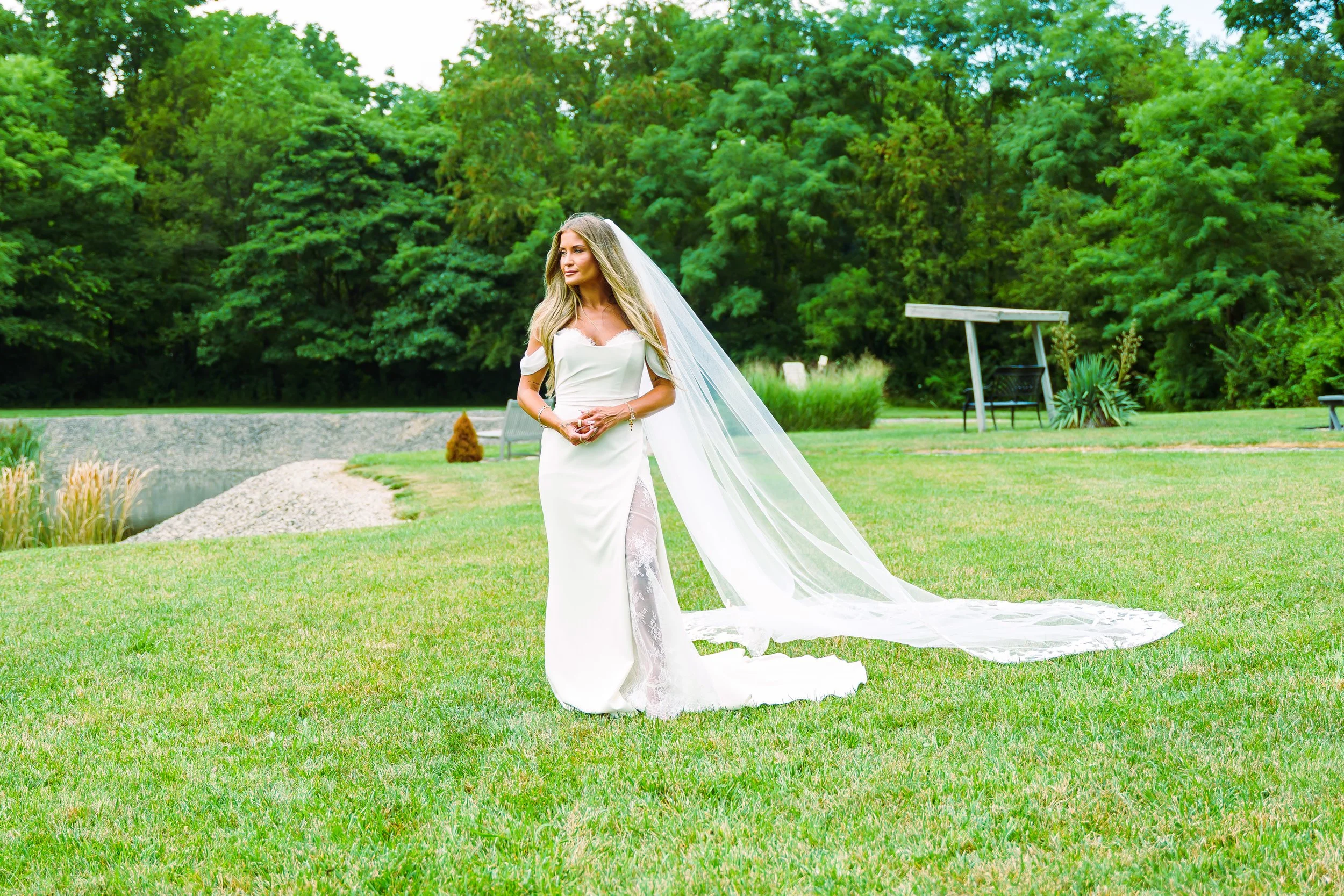 A bride in a white wedding gown with a long train and veil standing on a grassy lawn with lush green trees in the background.