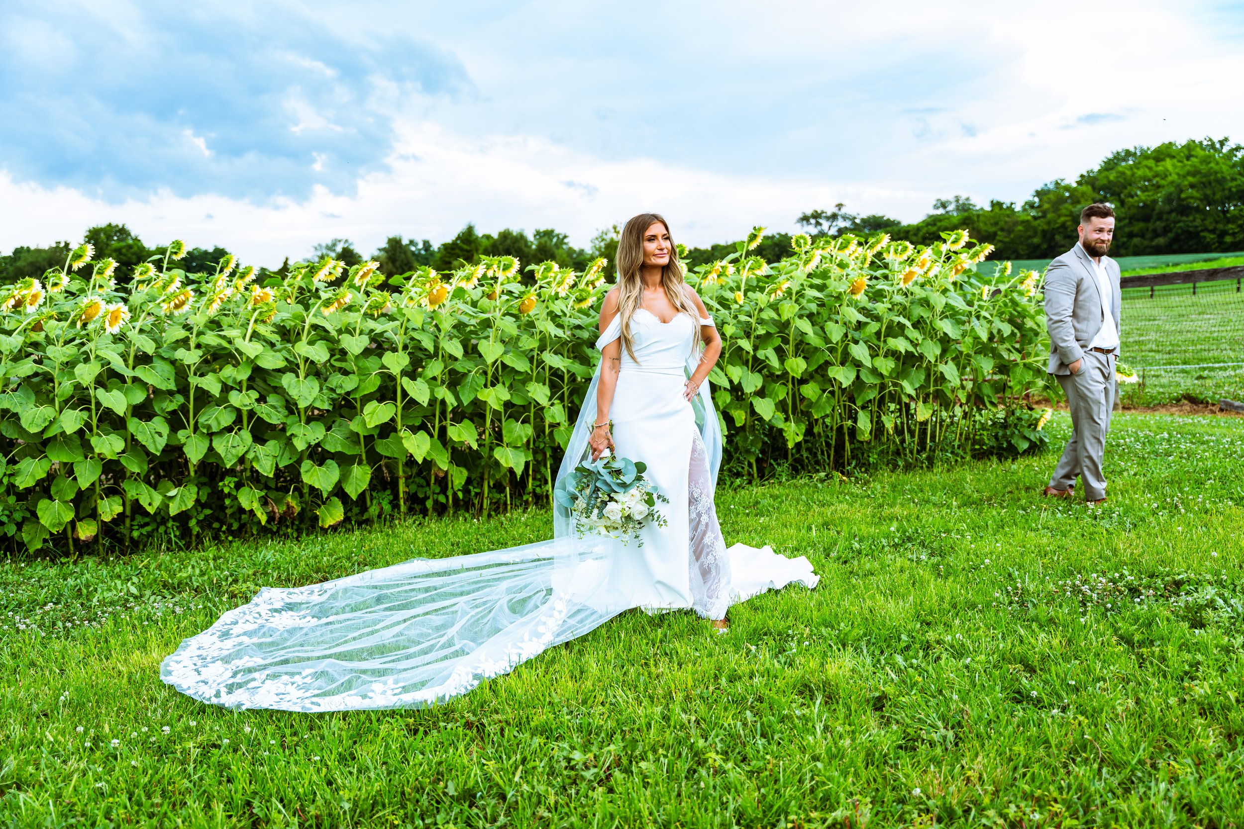 A bride in a white wedding dress holding a bouquet of flowers standing on a grassy field with sunflowers and a groom in a gray suit standing nearby, under a partly cloudy sky.