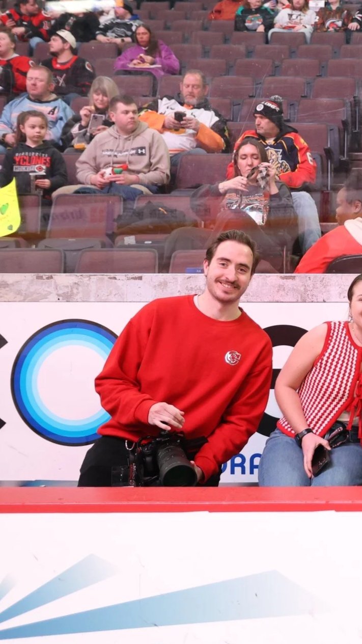 A smiling man in a red sweatshirt, holding a camera, is posing for a photo at a sporting event. Behind him, spectators are seated in the arena, some using their phones or watching the event.