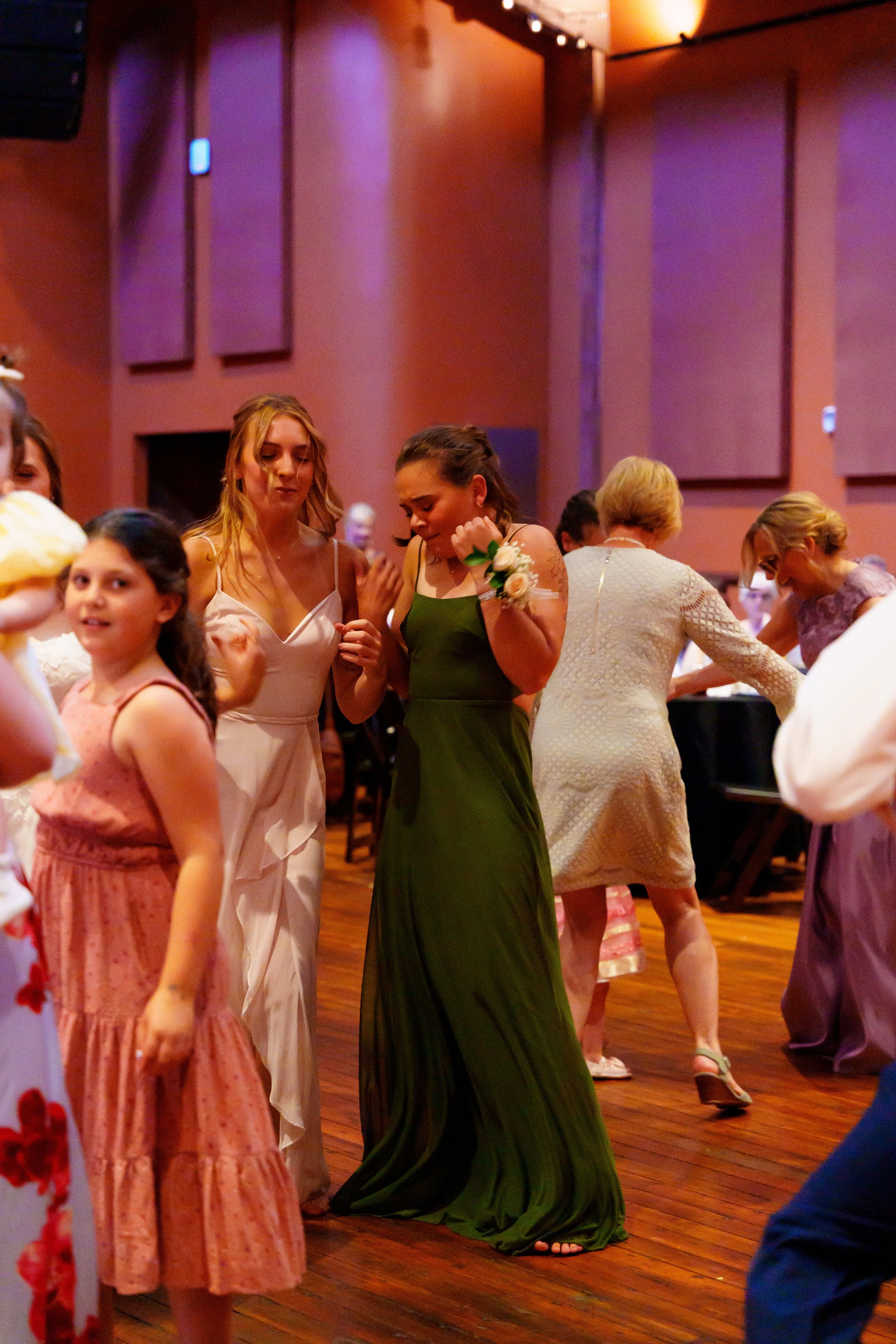 People dancing at a formal event or wedding reception in a decorated hall with wooden floors and warm lighting.