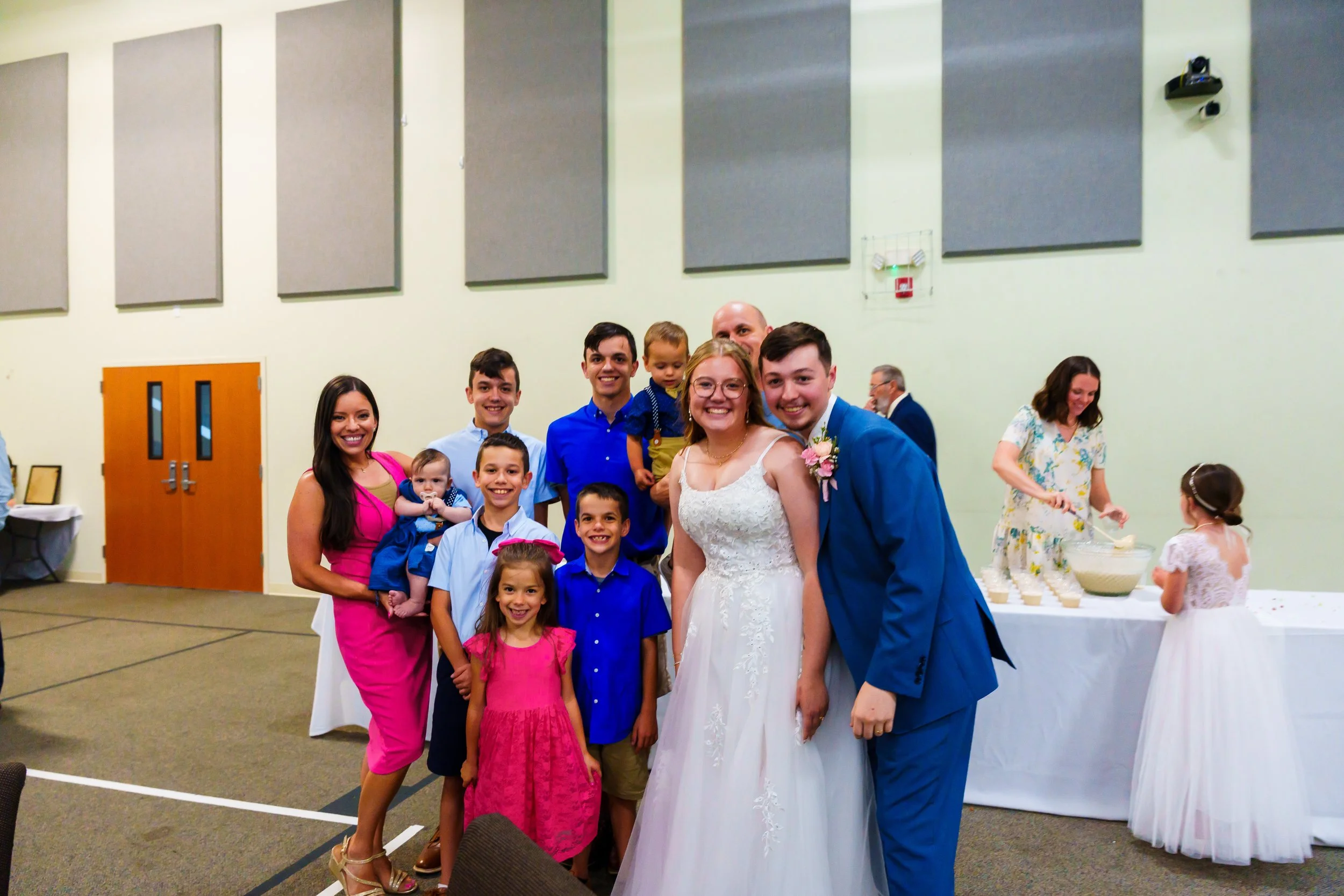 Group of people, including a bride and groom, smiling at a wedding reception in a large hall, with children and adults, and a table with cupcakes in the background.