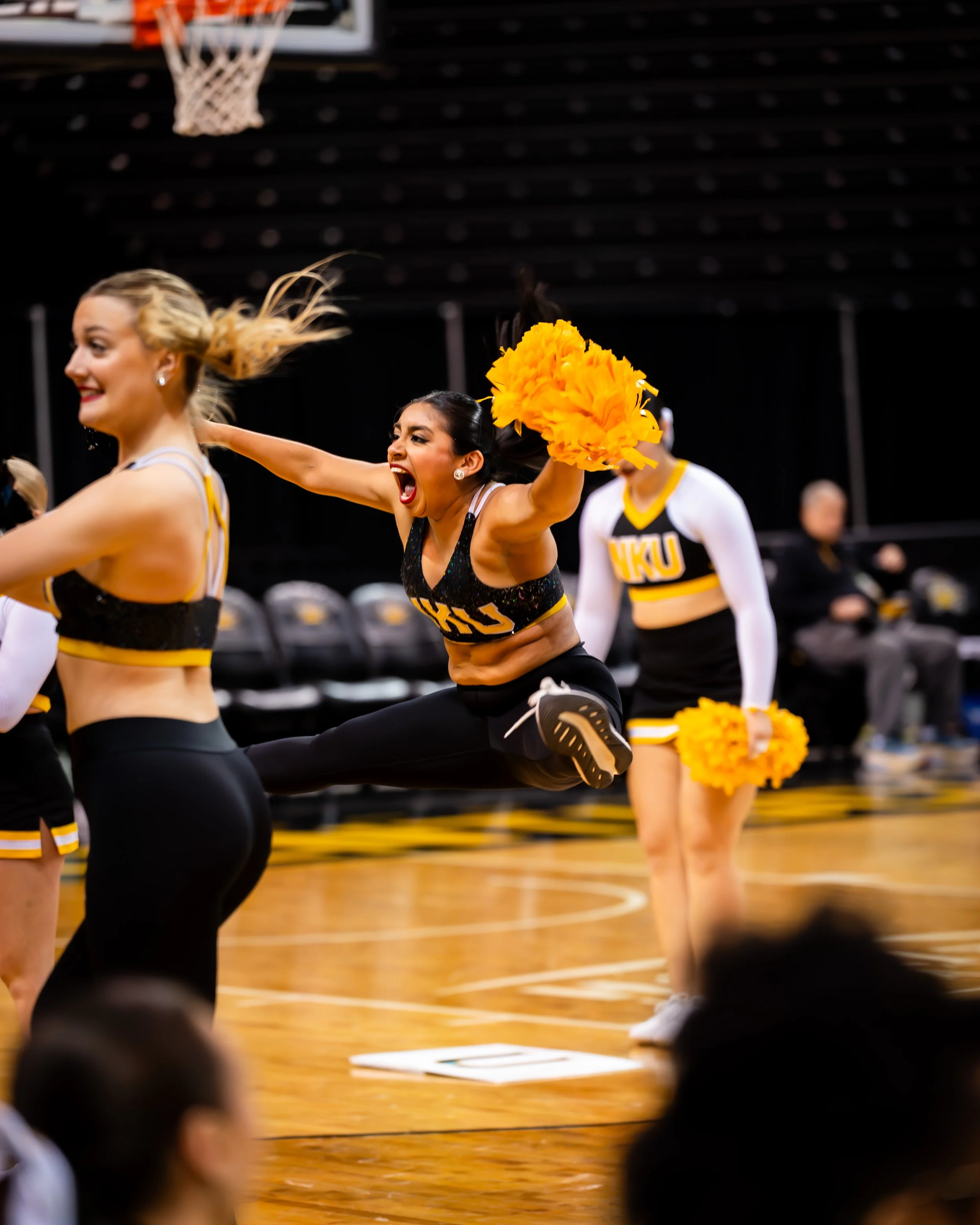 Cheerleaders performing a jump routine on a basketball court, wearing black and yellow uniforms with "NKU" lettering, holding orange pom-poms, with a black backdrop and seated audience.