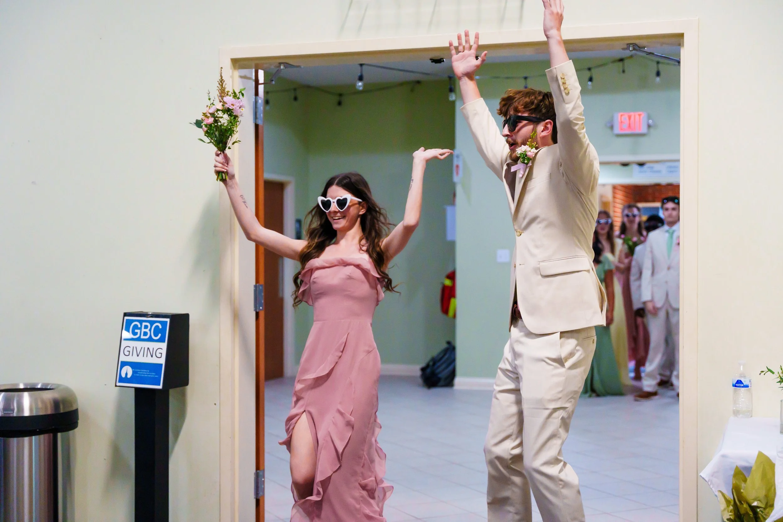 A woman in a pink dress wearing heart-shaped sunglasses holding a bouquet of flowers raises her arms, celebrating after an event. A man in a beige suit and sunglasses stands nearby with his arms up, smiling. People are visible in the background, some