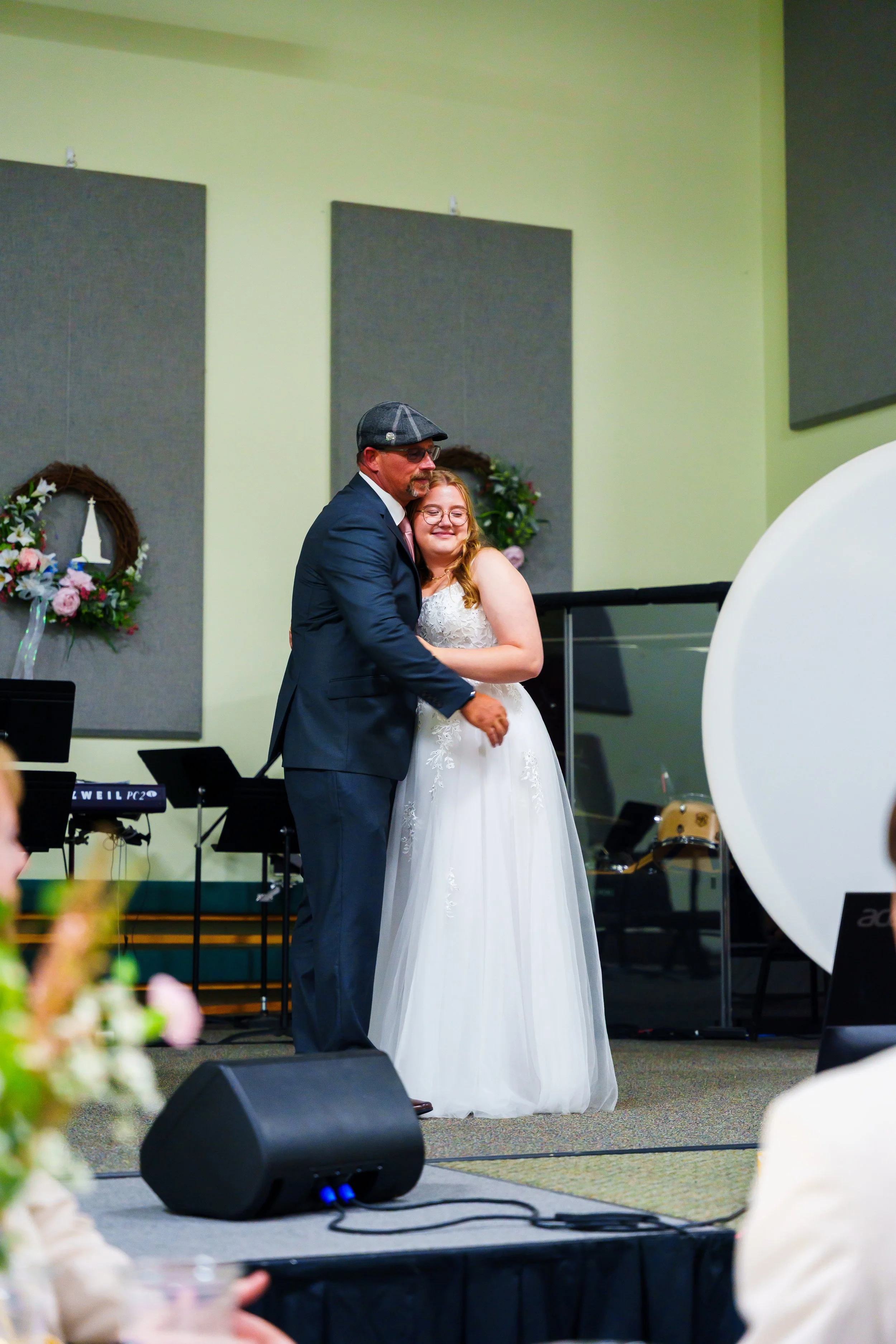 A man in a suit and cap dancing with a woman in a white wedding dress in a decorated hall.