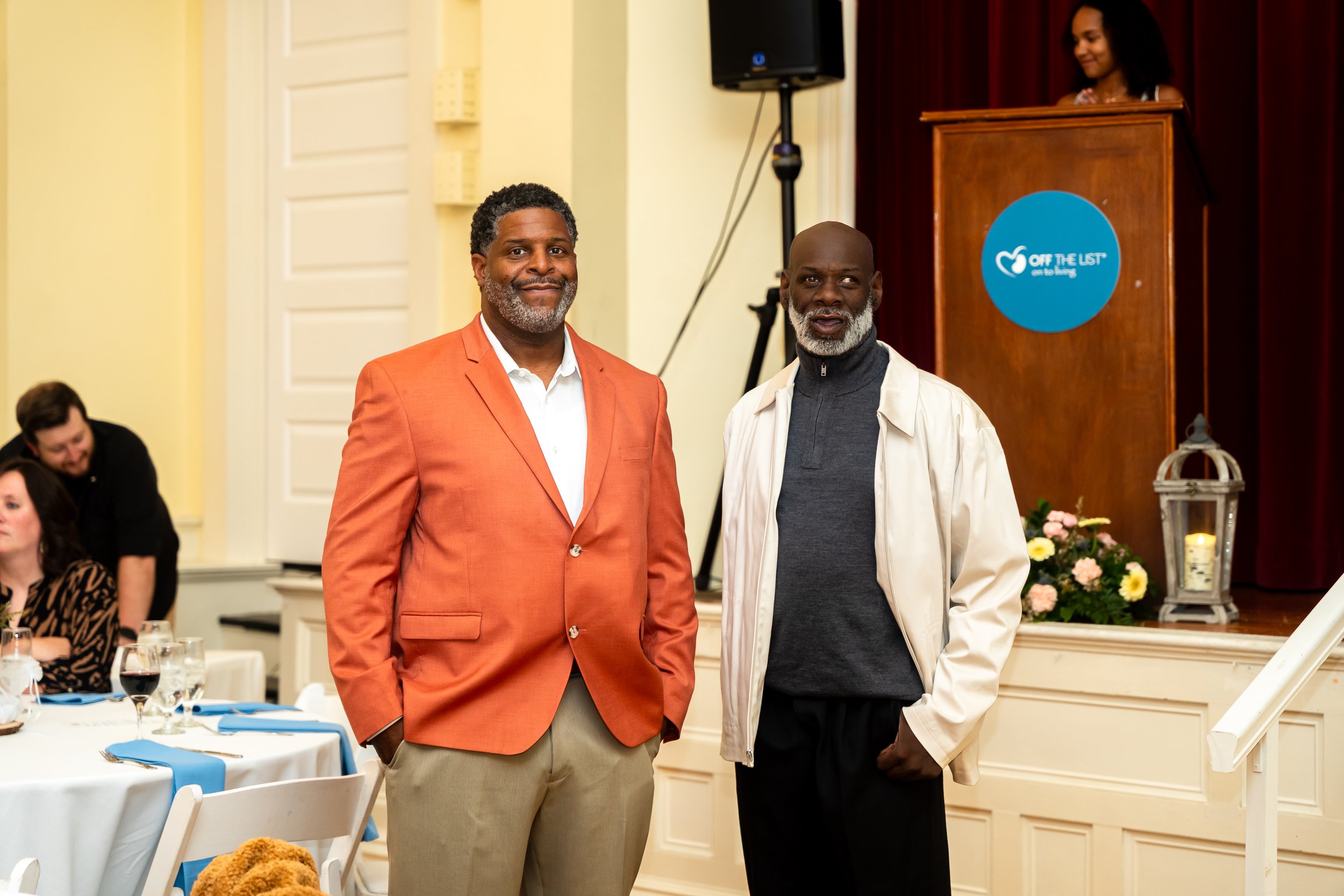 Two men standing together at a formal event, smiling at the camera. One wears a peach-colored blazer and the other wears a beige jacket with a gray shirt. In the background, other attendees are seated at tables, and a woman is standing at a podium wi