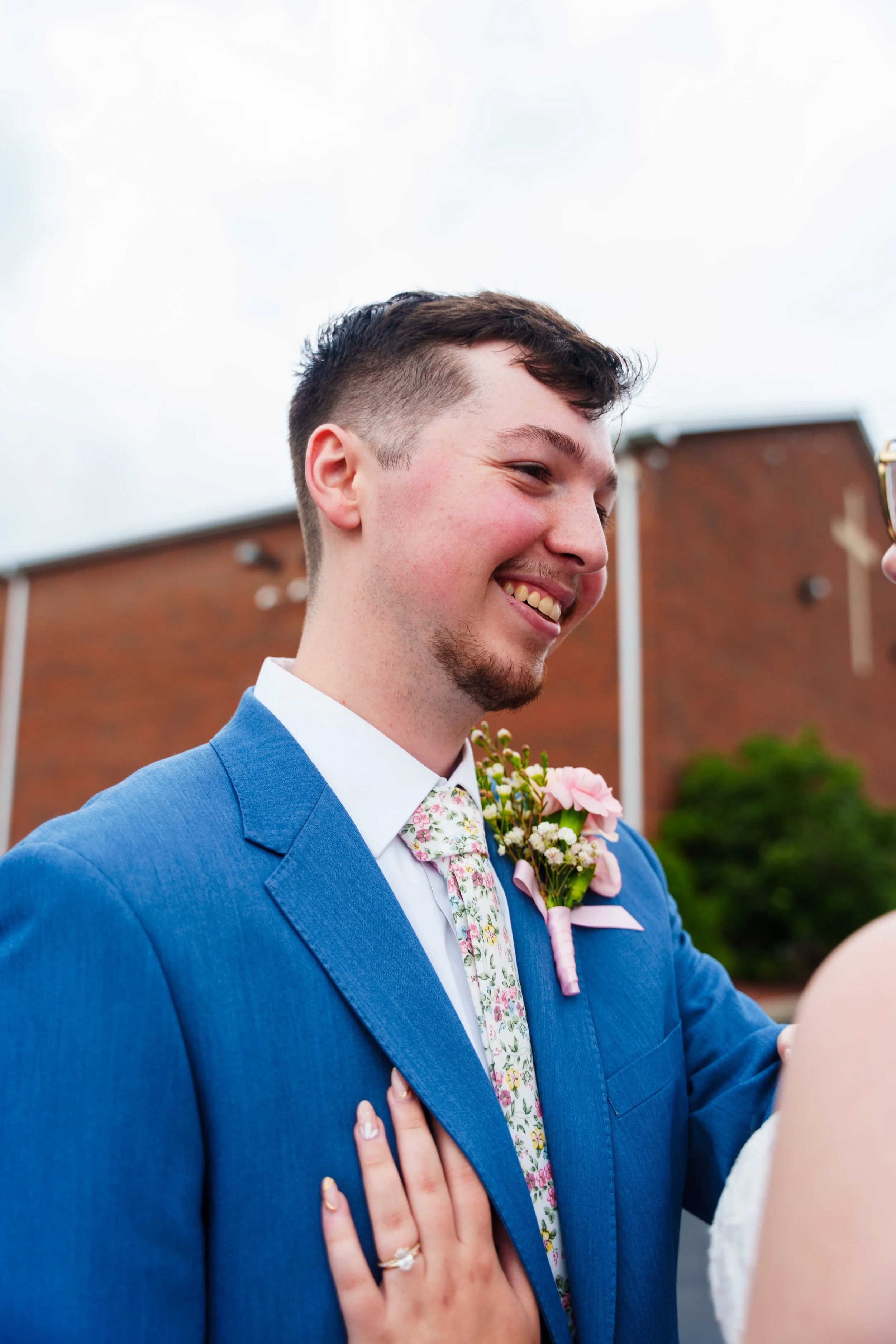 A smiling man in a blue suit with a floral tie and a pink boutonniere, likely at a wedding, with a woman touching his chest and wearing a ring.