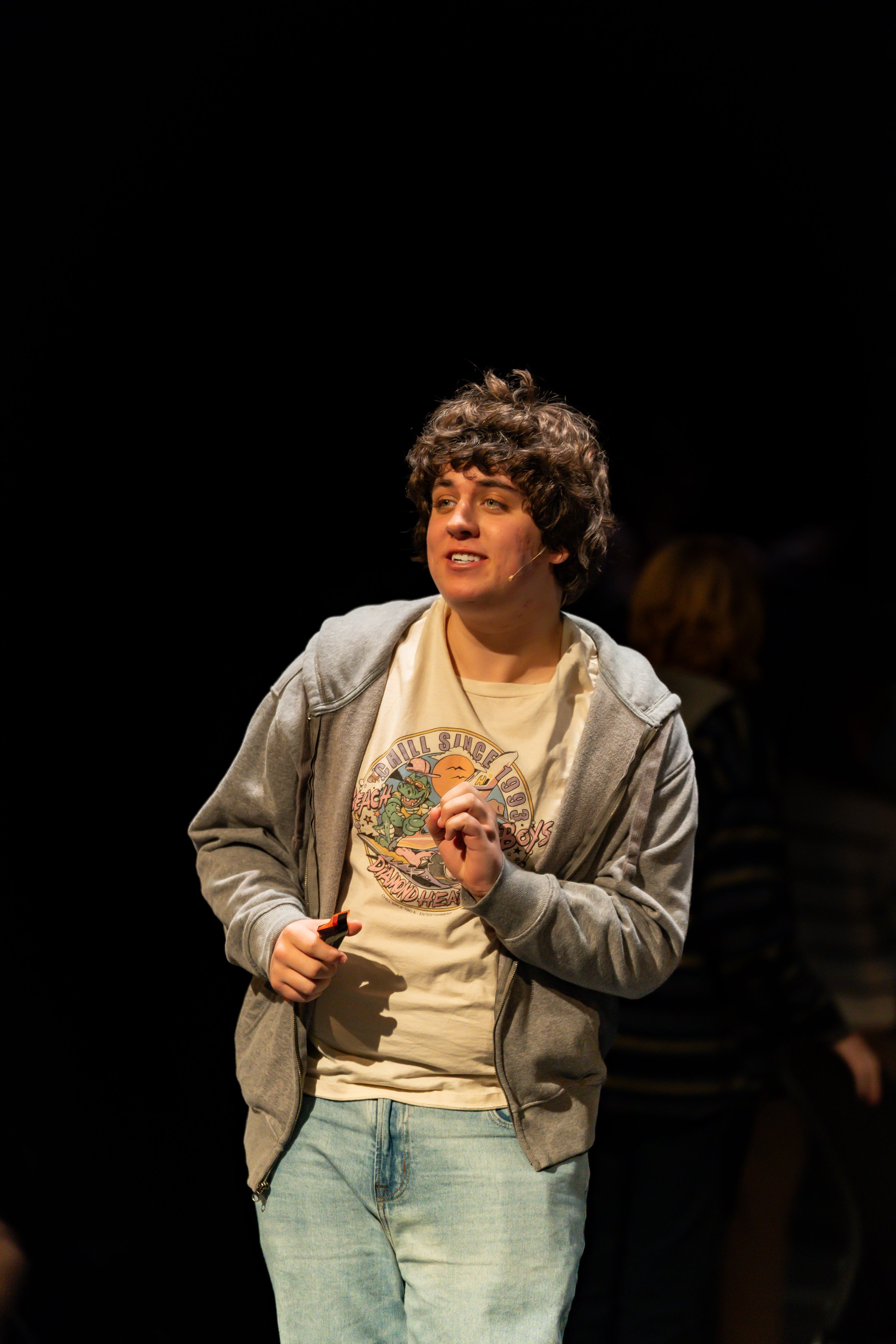 A young person with curly hair wearing a beige graphic t-shirt, light blue jeans, and a gray hoodie standing on a dark stage, speaking or performing, with a microphone headset.