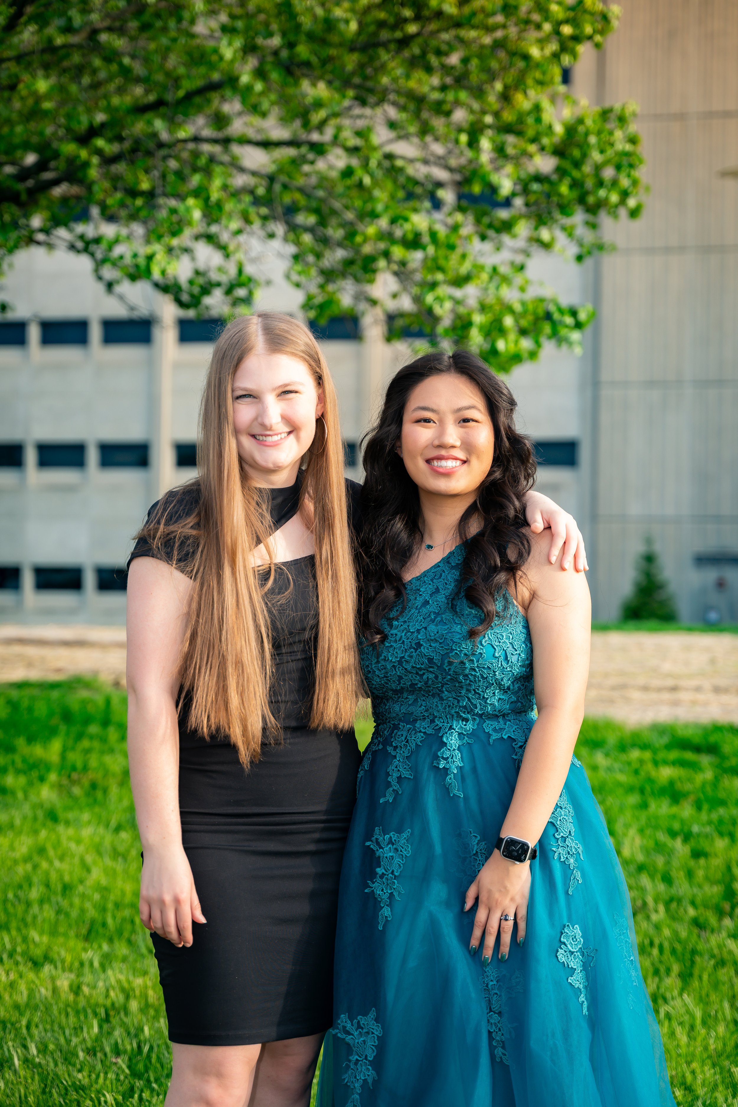 Two women standing outdoors, smiling, with a tree and a building in the background. One woman has long blonde hair, wearing a black dress, and the other has long dark hair, wearing a teal lace dress and an Apple Watch.