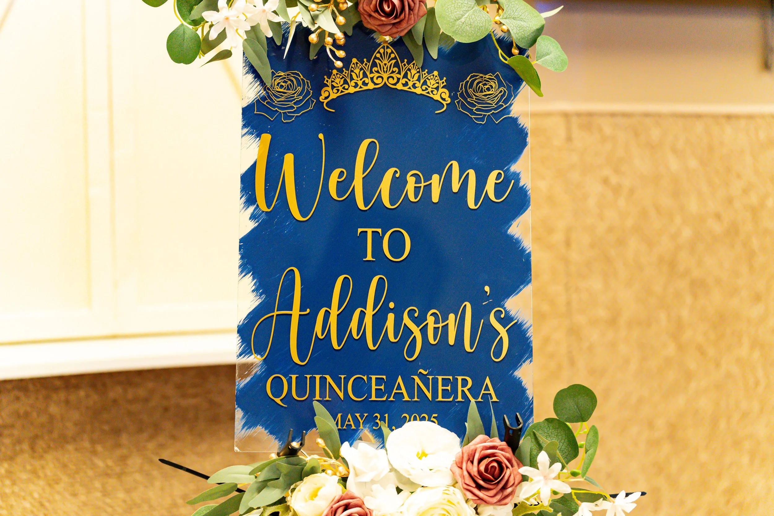 A floral arrangement with pink, white, and green flowers and leaves surrounding a blue vertical sign with gold text that reads "Welcome to Addison's Quinceañera" and the date "May 31, 2023." The sign is decorated with a gold tiara design at the top.