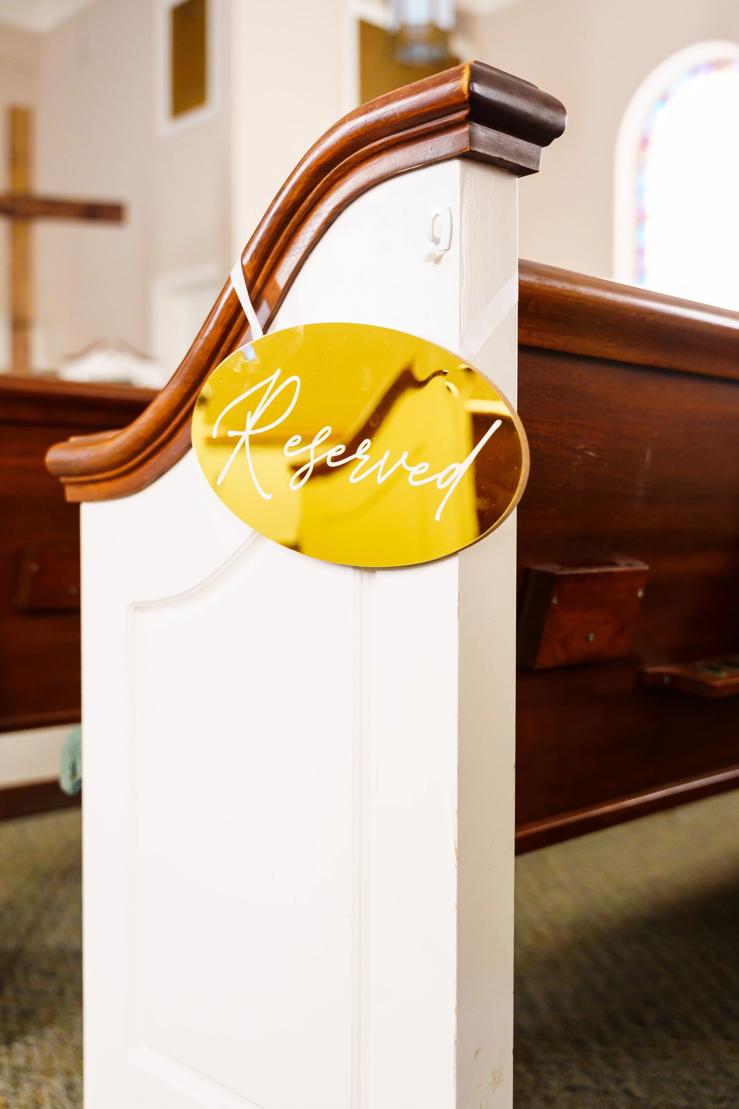 A white wooden church pew with a glossy brown wooden handrail on top, with a gold 'Reserved' sign hanging from it, in a church interior.