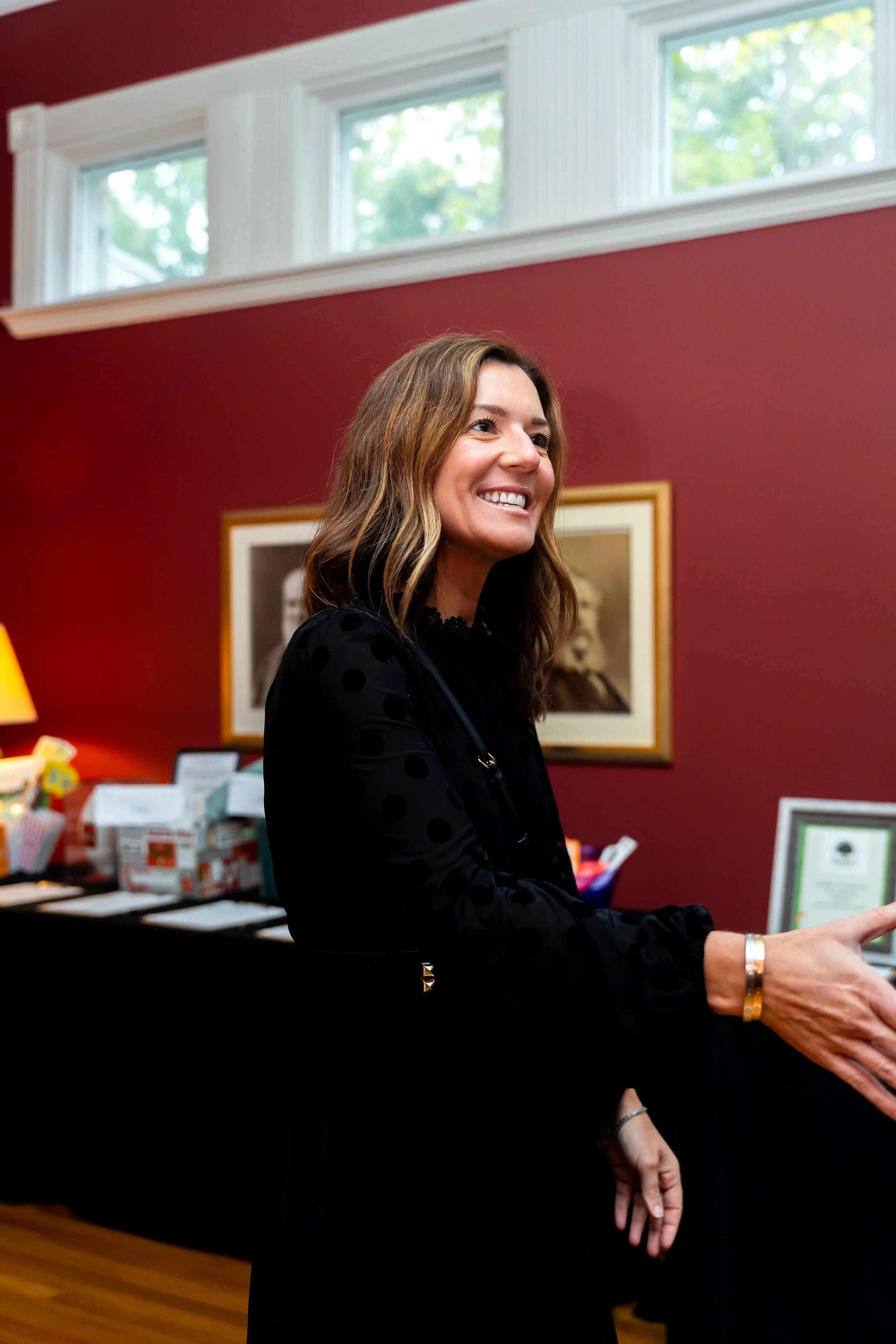 A woman with long wavy brown hair smiling, wearing a black polka-dot top, and standing indoors in front of a crimson wall with artwork framed on it, with windows above and a table with various items in the background.