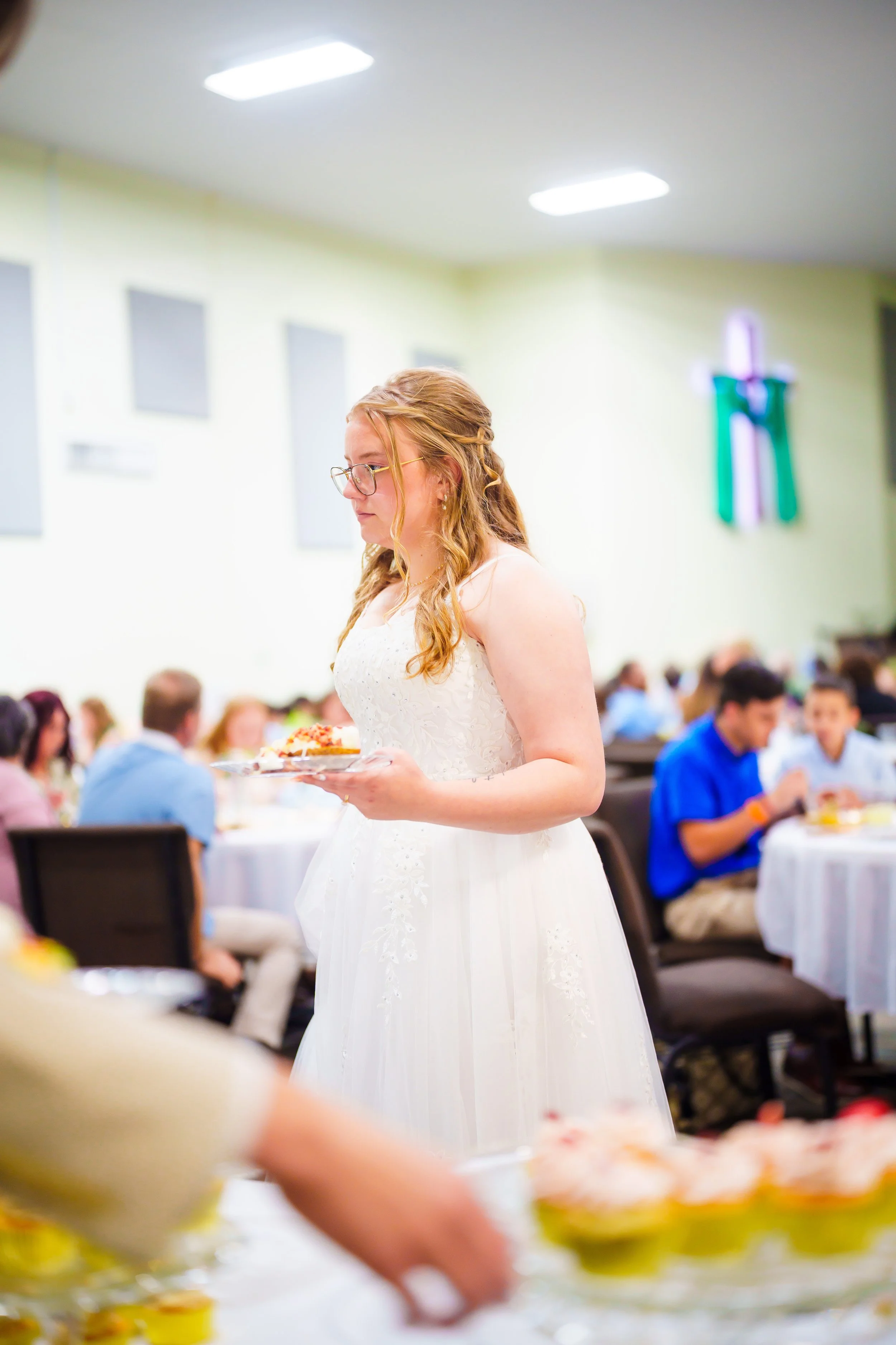 A young woman in a white dress and glasses holding a plate of food at a gathering in a hall with people seated at tables in the background.
