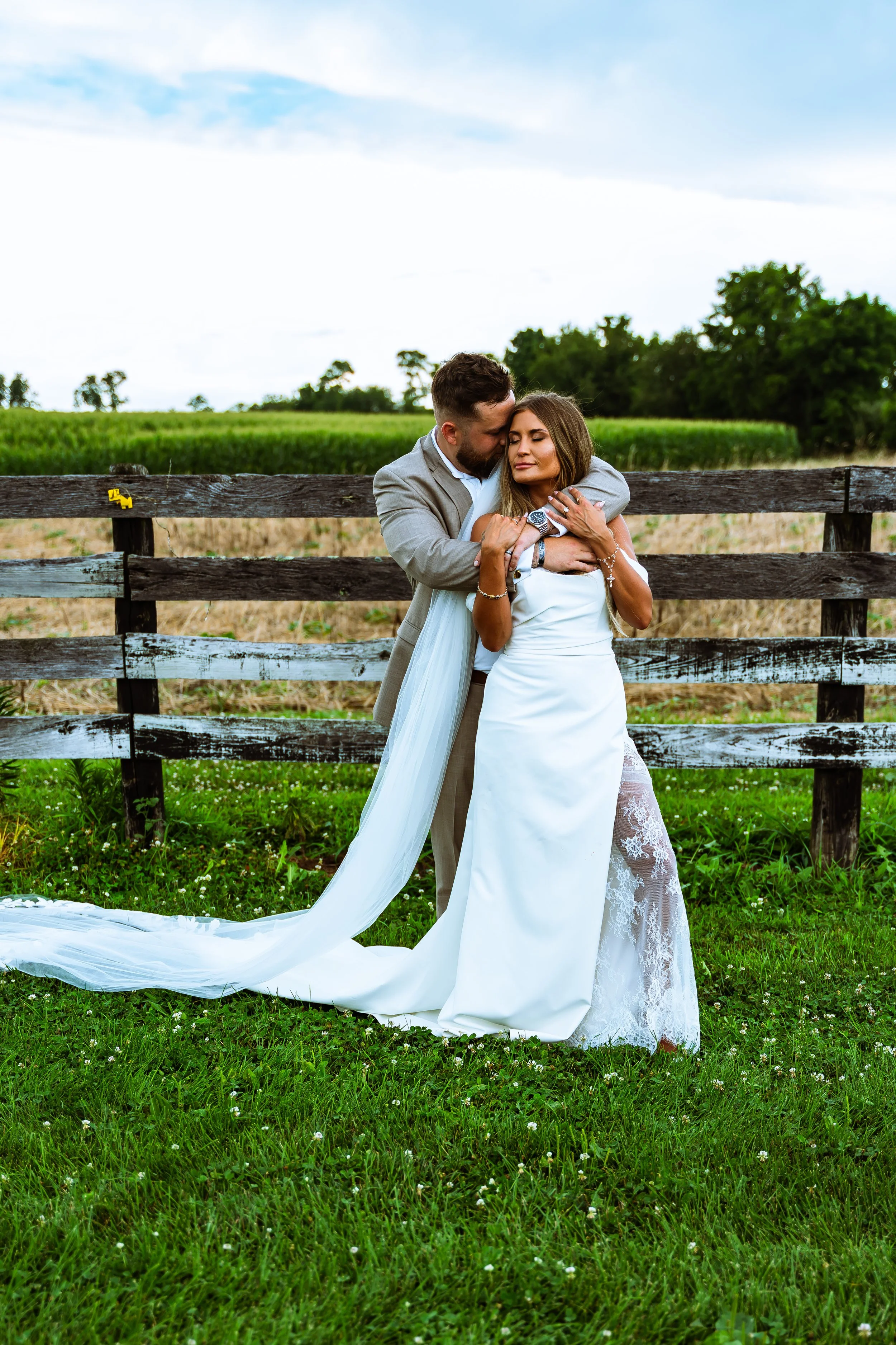 A newlywed couple embracing and kissing outdoors in a lush green field with a rustic wooden fence, trees, and a partly cloudy sky in the background.