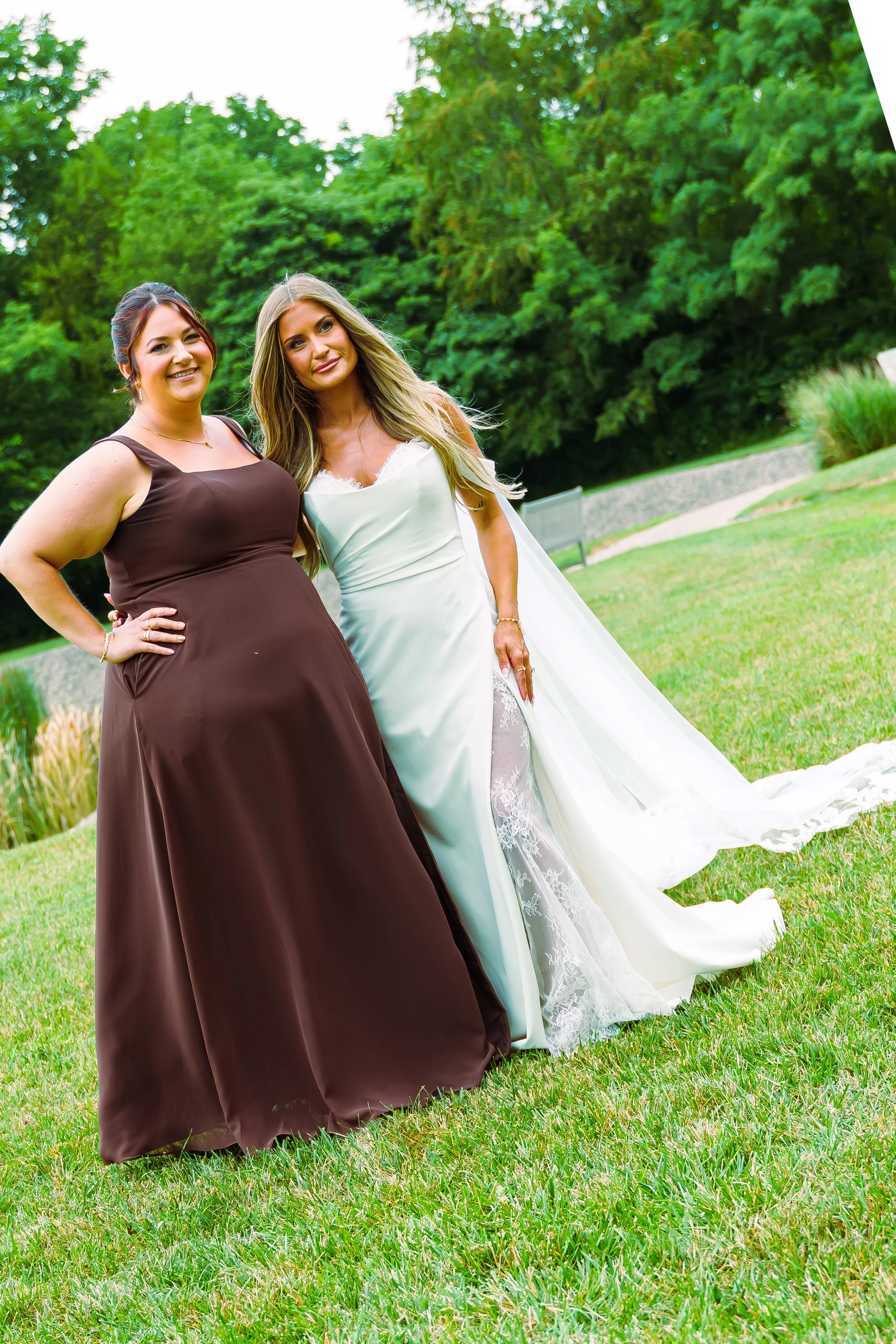 Two women standing outdoors on a grassy lawn with trees in the background, one wearing a brown dress and the other in a white dress, smiling at the camera.
