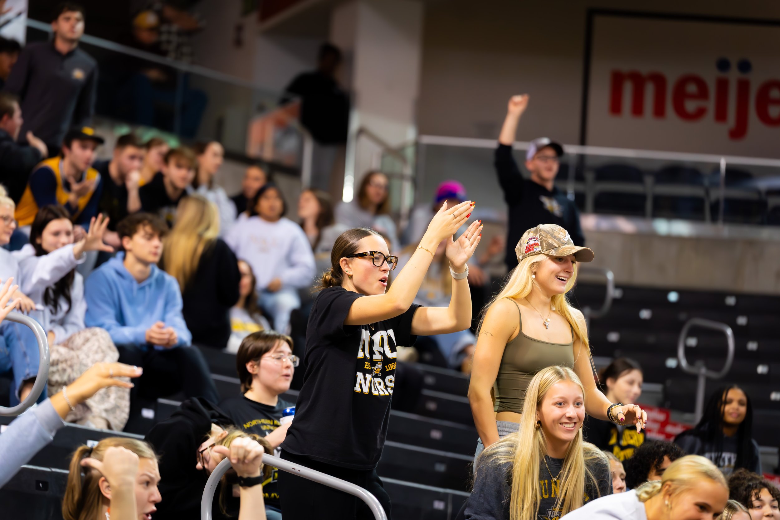Cheering crowd of mostly young women and girls at a sports event, some standing and clapping, with cheering and excitement visible.