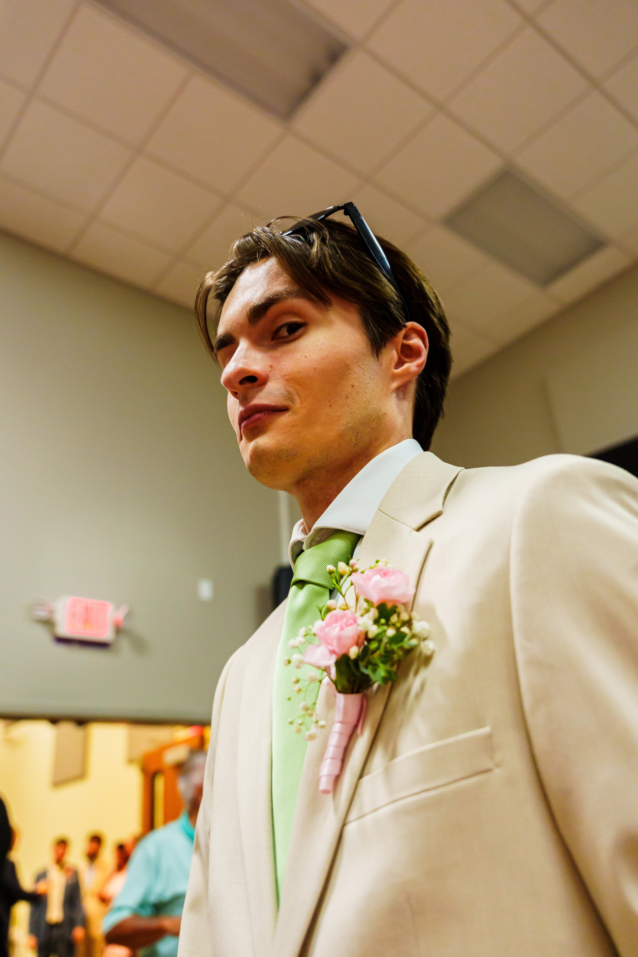 A young man in a beige suit with a light green tie and pink flower boutonniere, looking over his shoulder at a wedding or formal event, with a beige ceiling and hallway in the background.