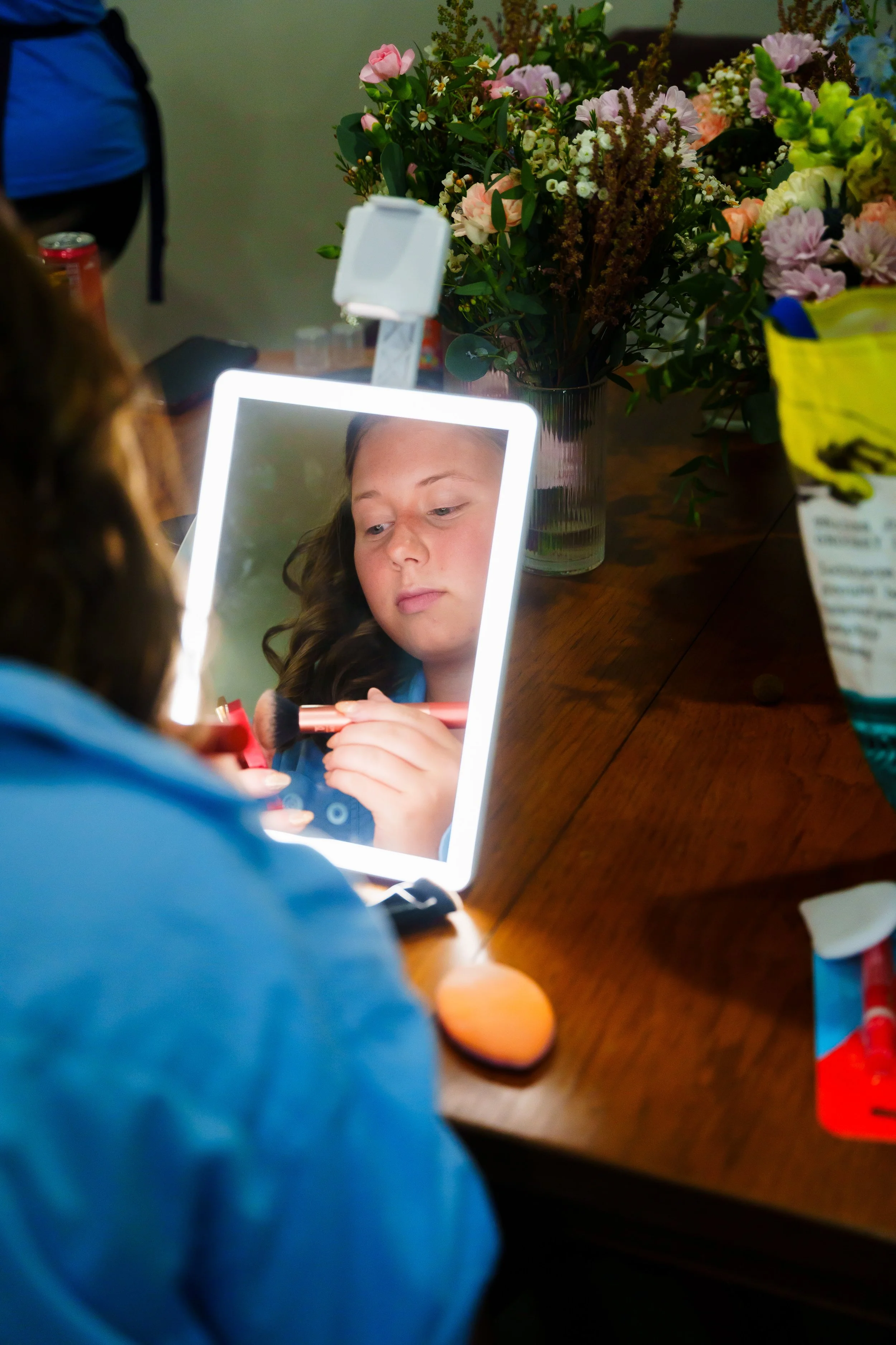 A person with curly hair is applying makeup in front of a mirror with a bright LED border. The person is holding a makeup brush and the reflection shows their focused expression. In the background, there is a table with a large bouquet of mixed pink,