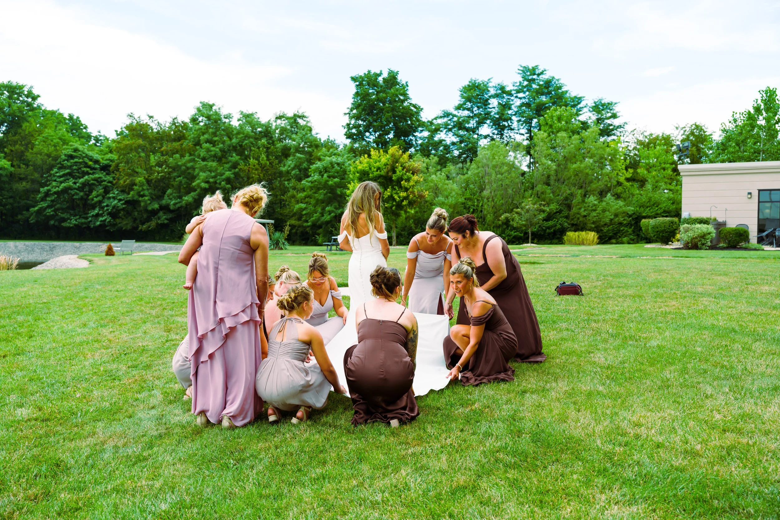 A group of women, dressed in formal gowns, participating in a wedding ceremony outdoors on a grassy area, with trees and a building in the background.
