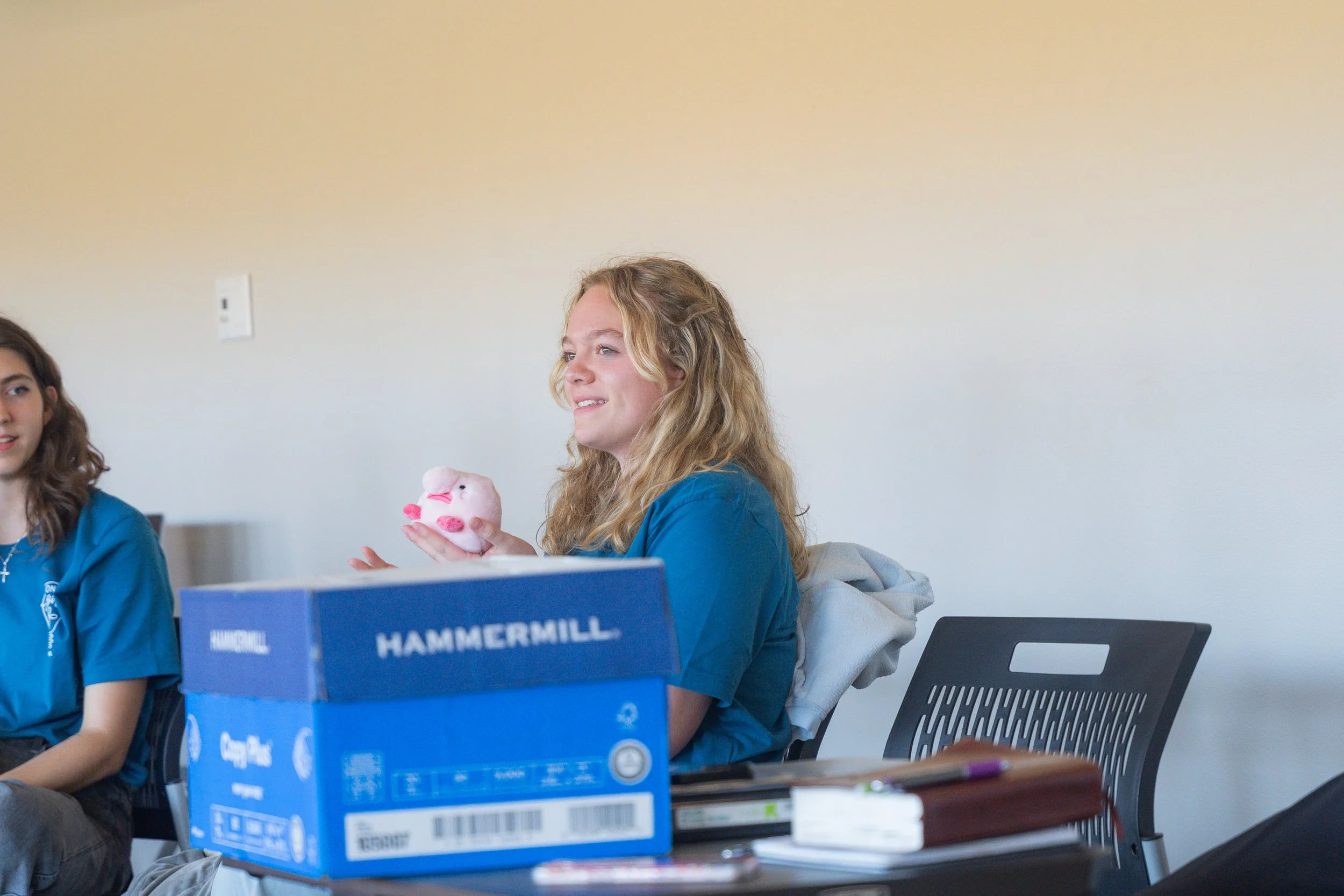 A young woman with curly blonde hair and a blue shirt sitting at a table in a room with plain white walls, holding a pink plush toy and smiling. There is a blue box labeled 'HAMMERMILL' on the table, along with other items including a notepad and som