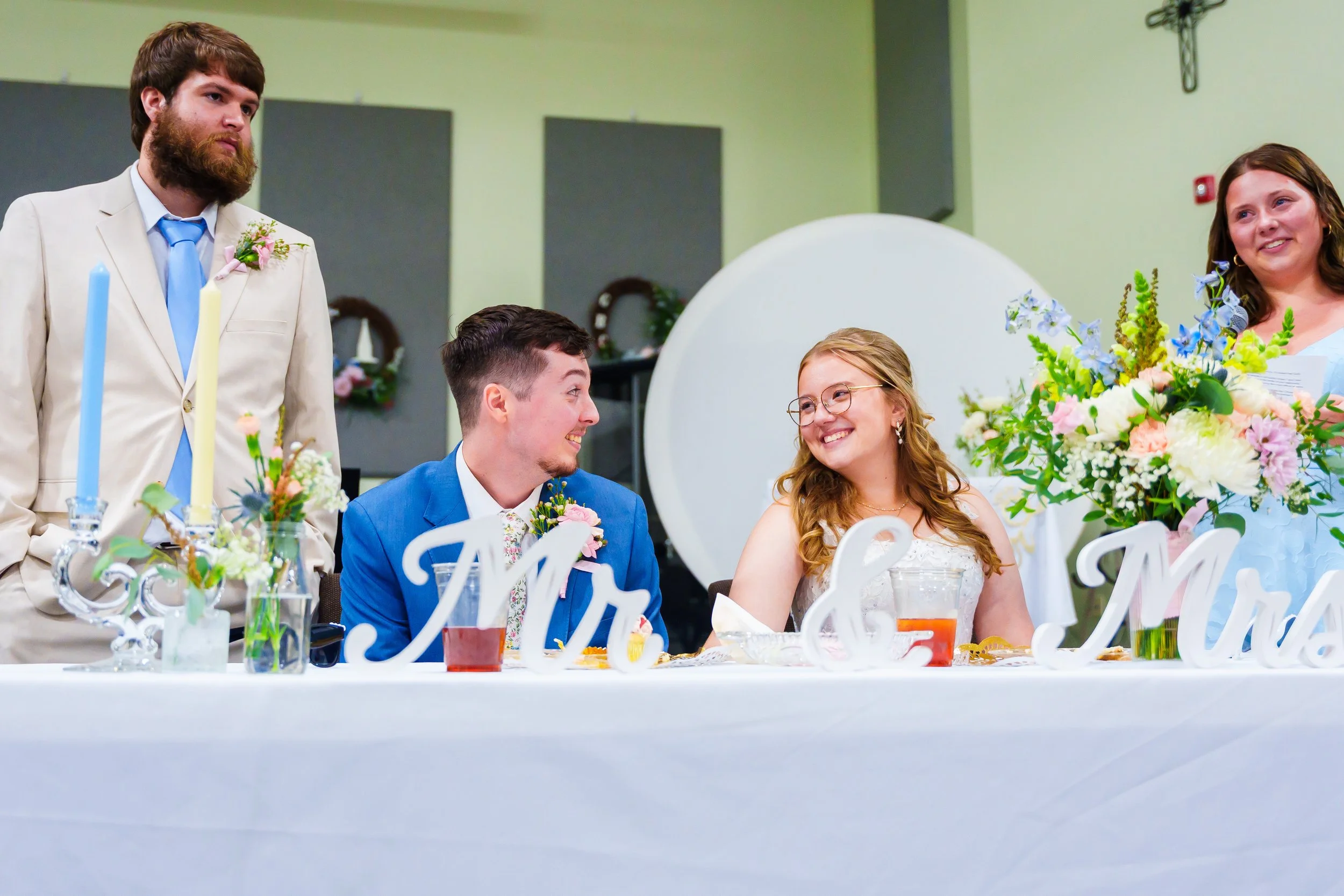 A bride and groom seated at their wedding reception table, surrounded by guests, with floral arrangements and candles, in a decorated indoor venue.