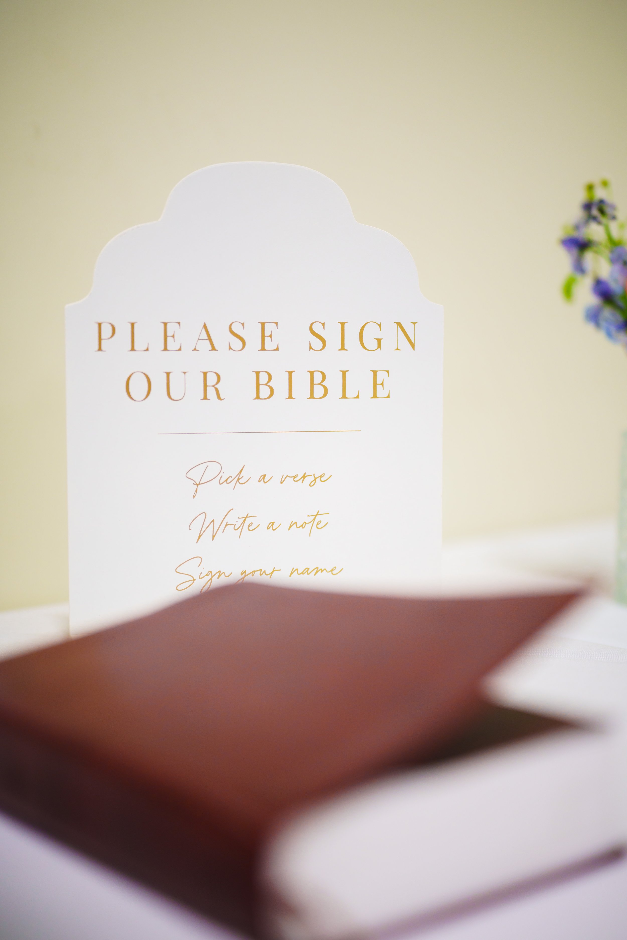 A sign on a church wall with the words "Please Sign Our Bible" and instructions to pick a verse, write a note, and sign your name, with a blurred hand in the foreground.