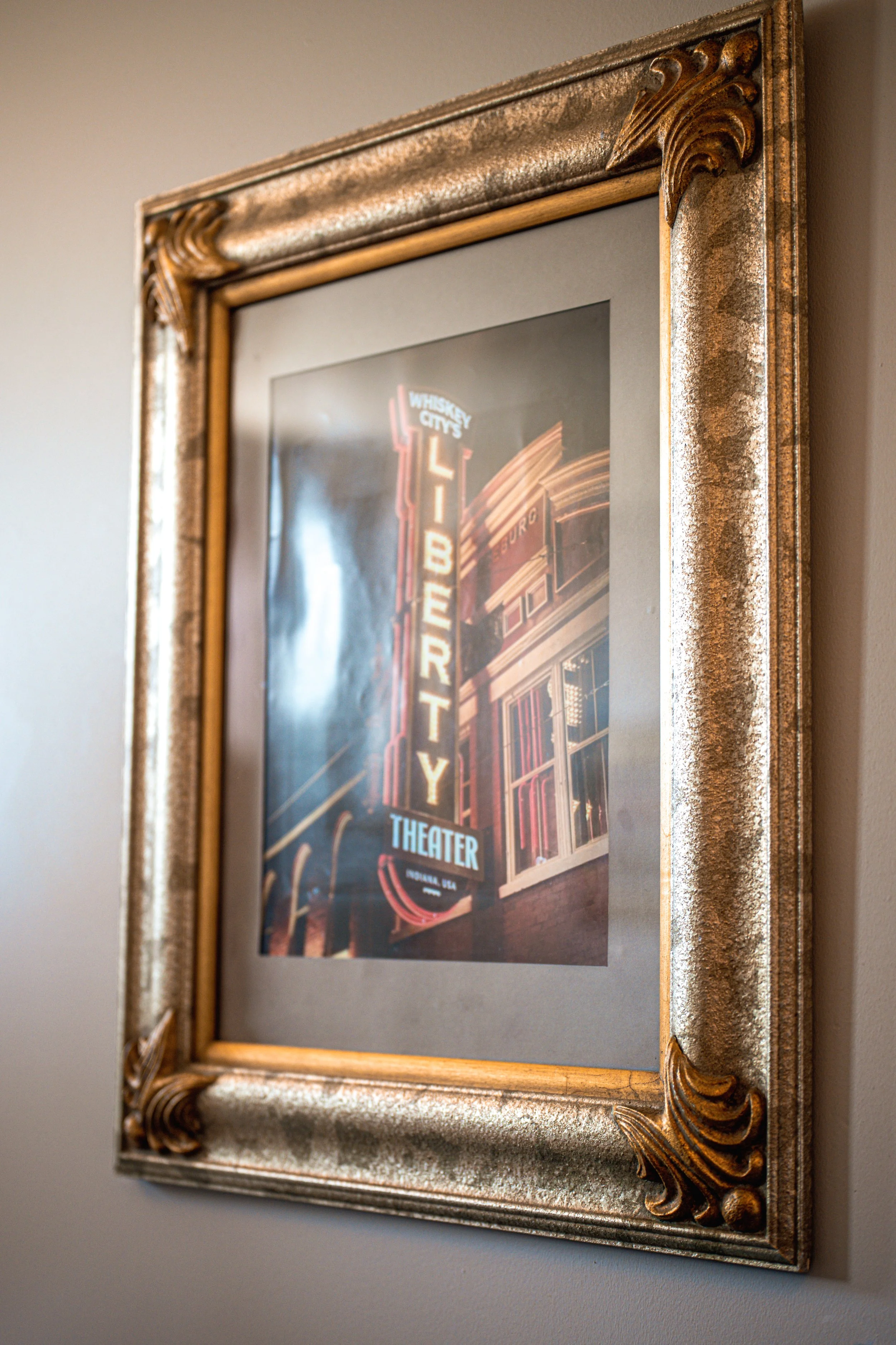 Close-up photo of a framed poster artwork on a wall, featuring neon sign art for Whiskey City Theater, with warm lighting highlighting the ornate gold and silver frame.