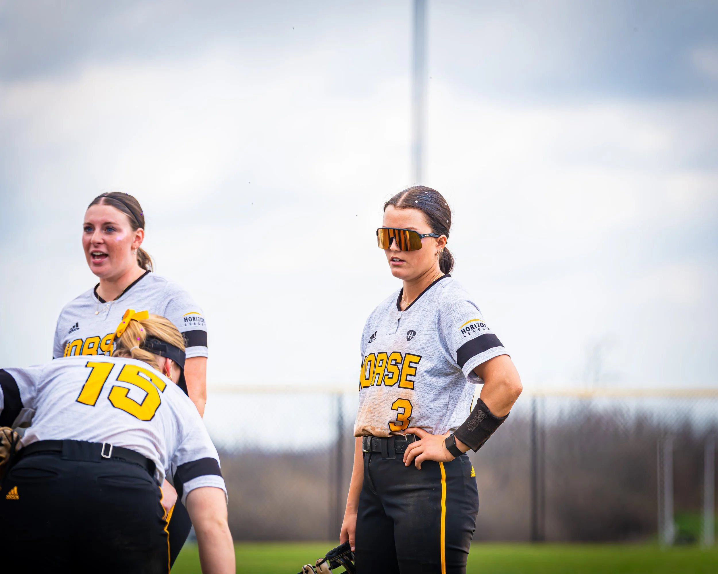 Three female softball players standing on a field, wearing gray uniforms with yellow and black accents, with a cloudy sky in the background.