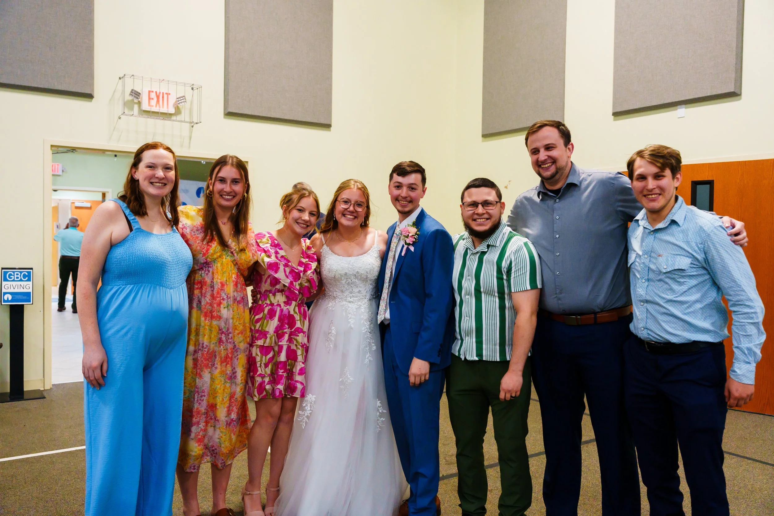 Group of eight people smiling and posing together at an indoor event, with a woman in a white wedding dress at the center.