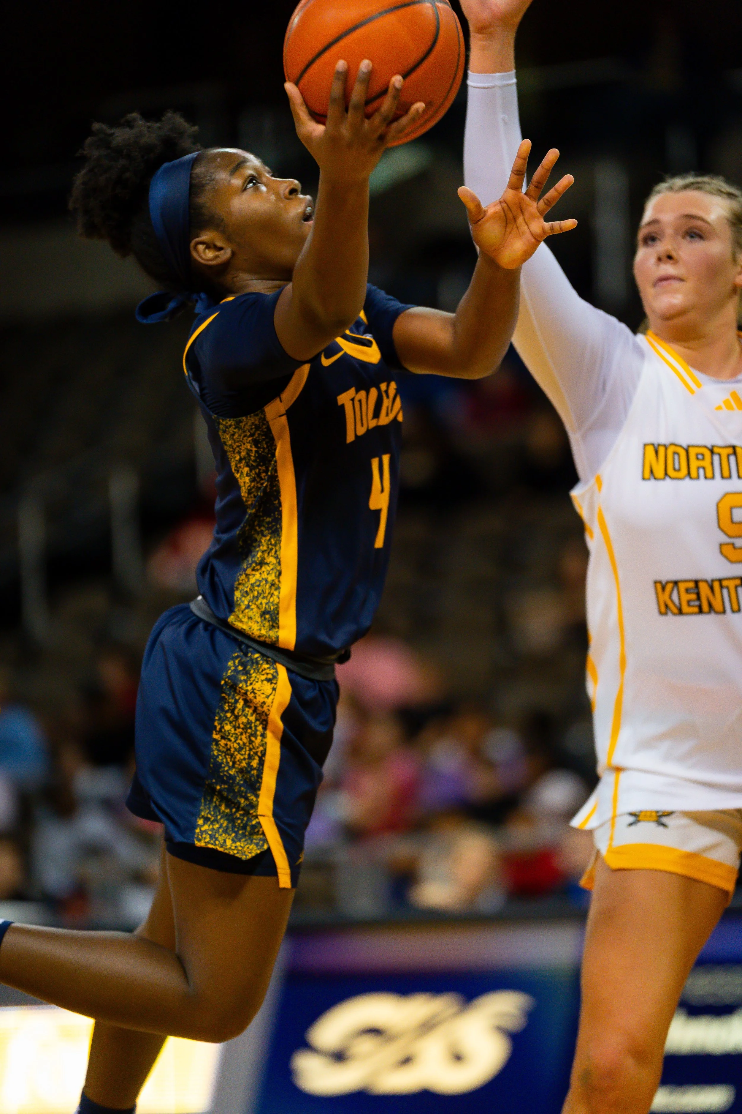 A female basketball player from Toledo attempting a shot, being guarded by a female player from a team with North Kentucky jersey in an indoor basketball game.