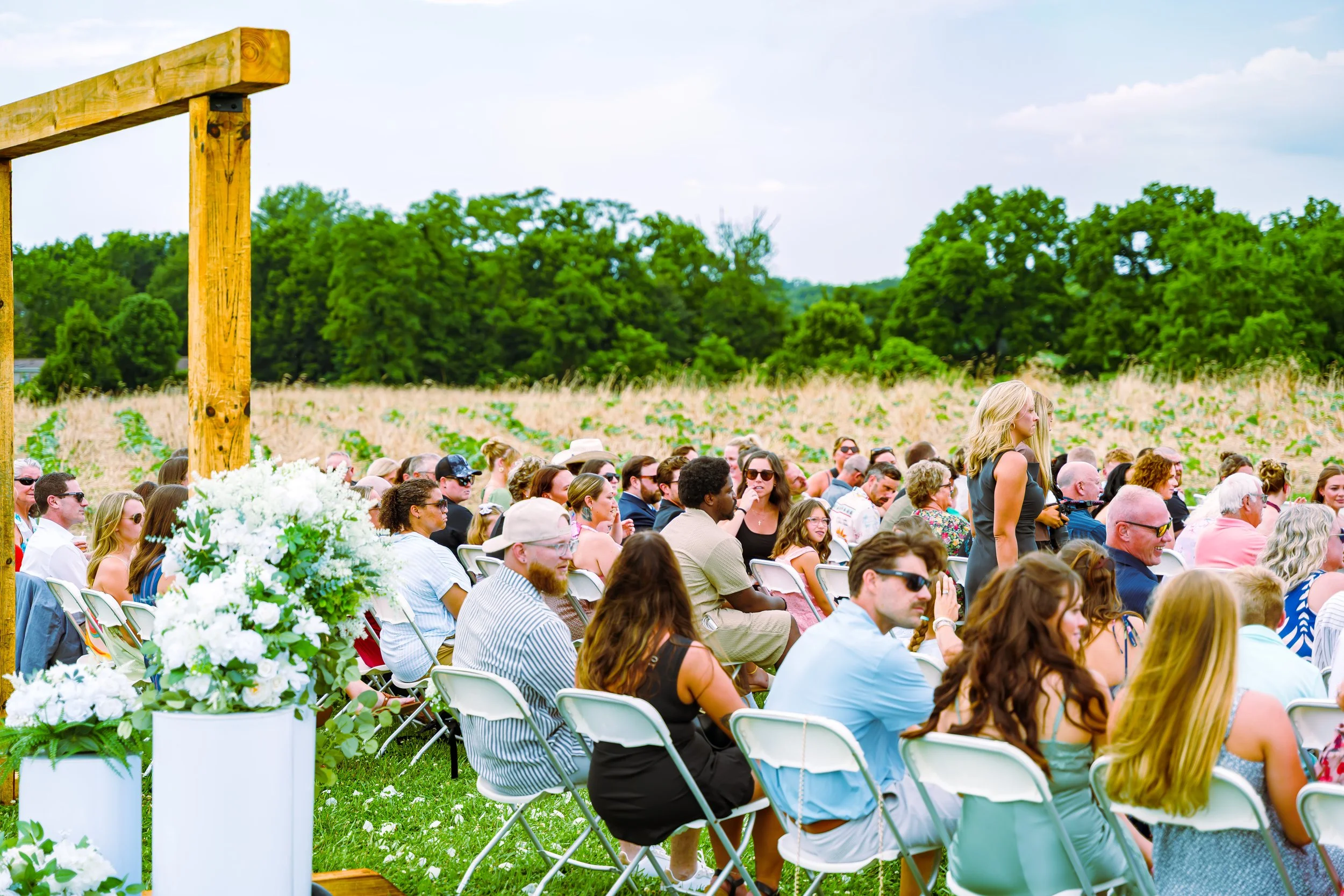 Outdoor event with many people sitting on chairs, some standing, in front of a green field with trees, under a partly cloudy sky.