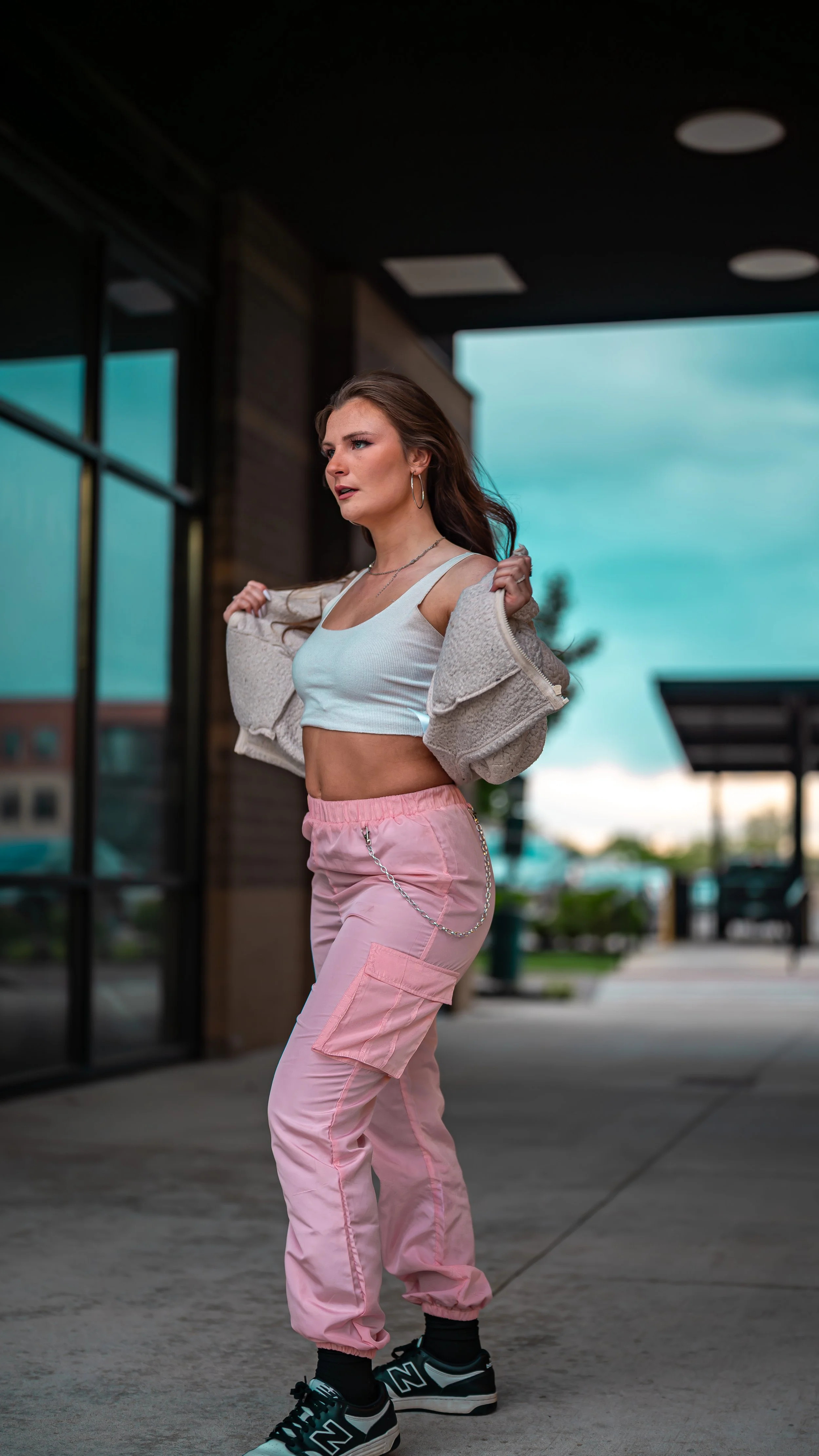 Young woman standing outside near a building with large glass windows, wearing a white crop top, beige jacket, pink cargo pants with chains, black sneakers, and hoop earrings, looking to the side.