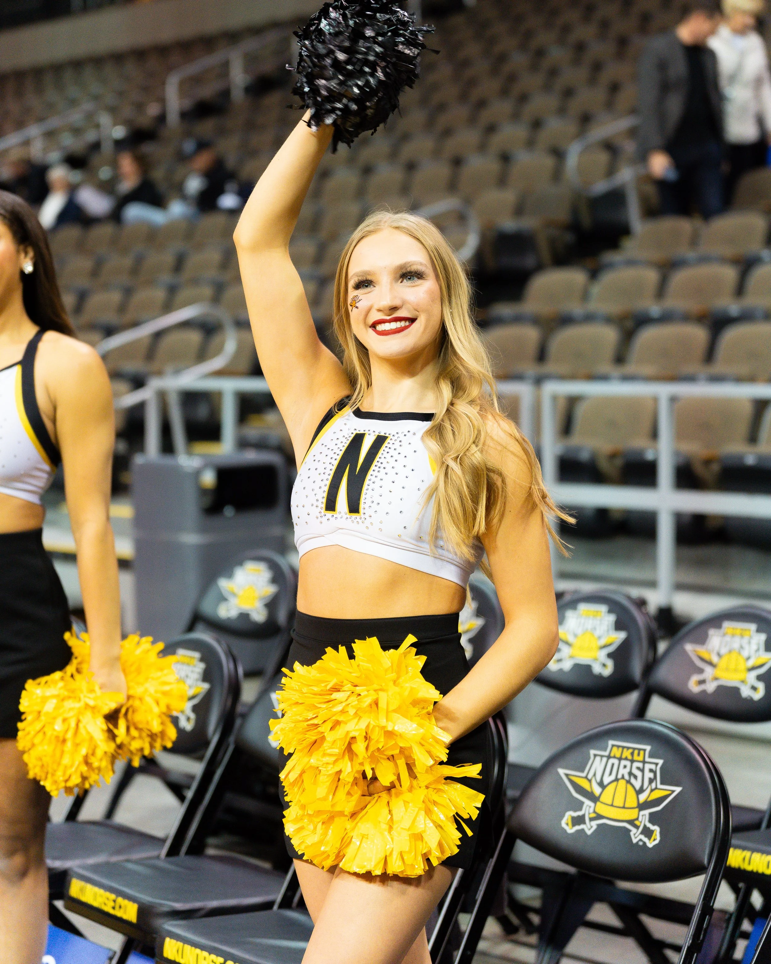 A cheerleader with a blonde ponytail, wearing a white and black uniform with the letter 'N' on it, smiling and holding yellow pom-poms while raising one arm in a sports arena with empty seating.