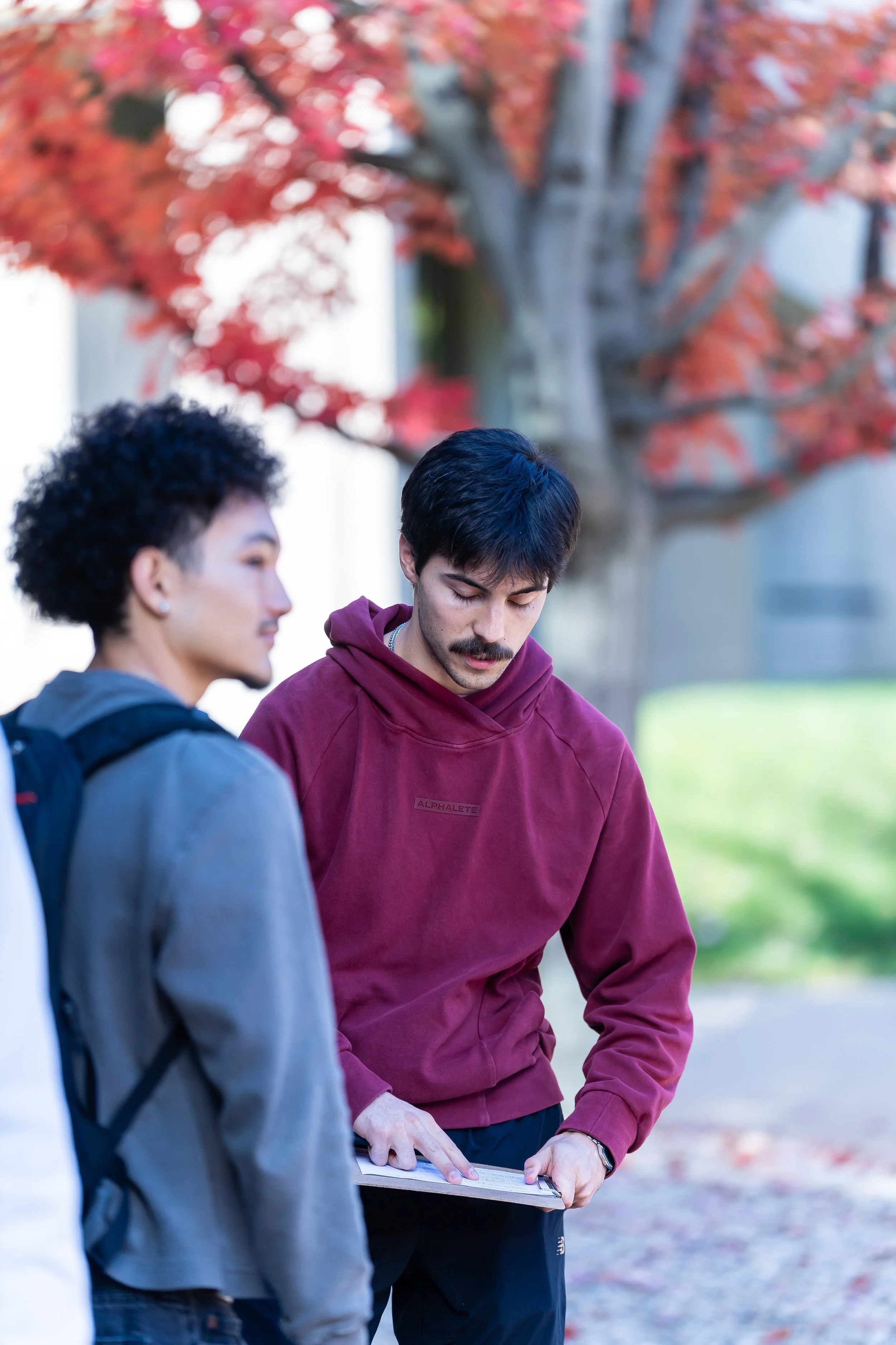 Two men standing outdoors under a tree with red fall foliage, one is holding a clipboard and writing, the other is listening and looking down at the clipboard.