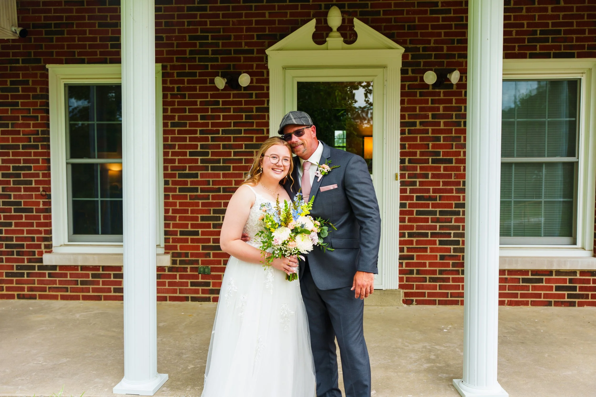Bride in a white wedding dress holding a bouquet of flowers standing next to groom in a suit with a pink tie and a hat, outdoors in front of a brick house with white trim and windows.
