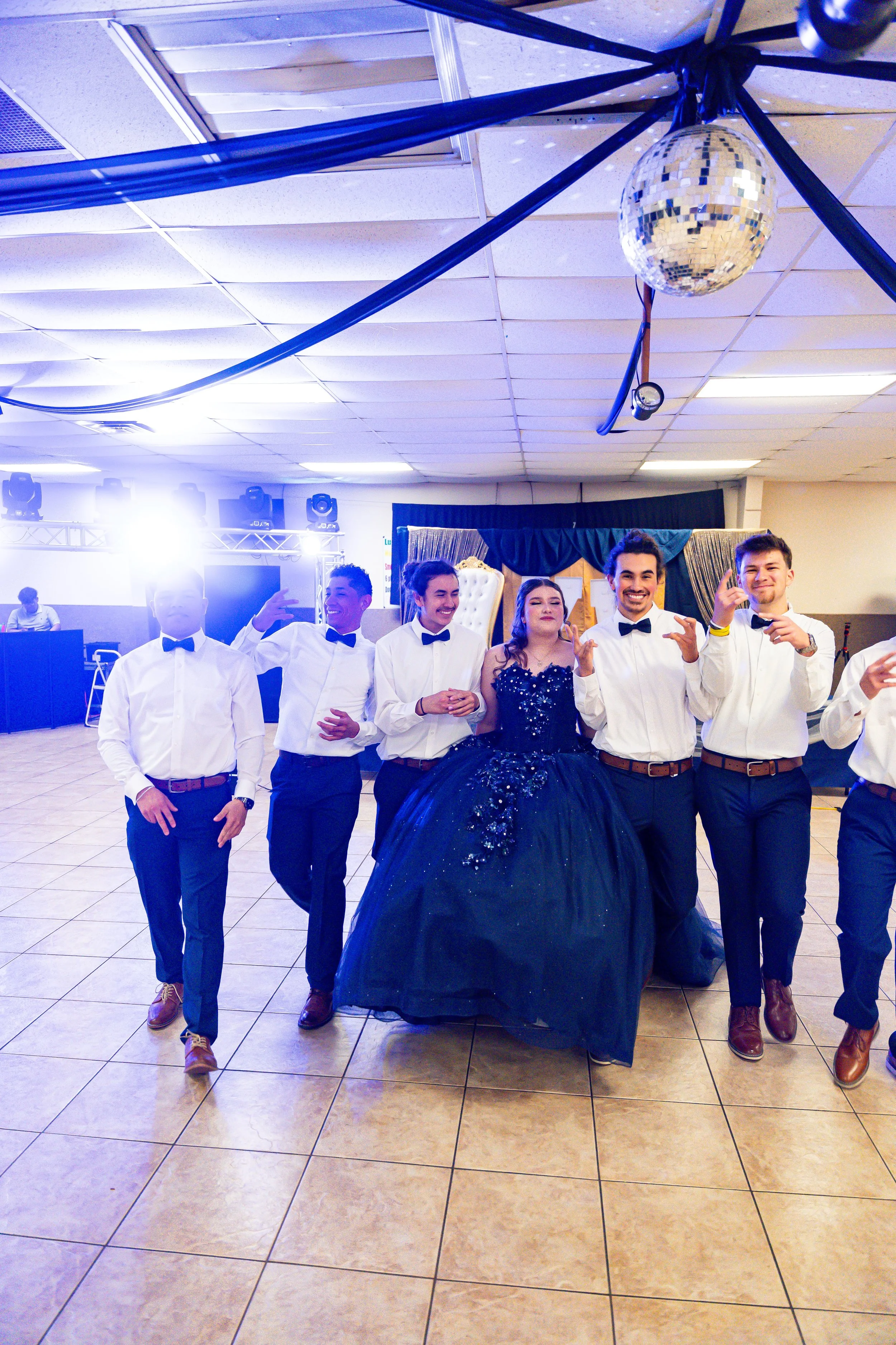A group of young people dressed in formal attire standing together at a party or celebration, with some wearing white shirts, black bow ties, and dark pants, and a young woman in a dark, sparkling gown, smiling and posing for a photo on a dance floor