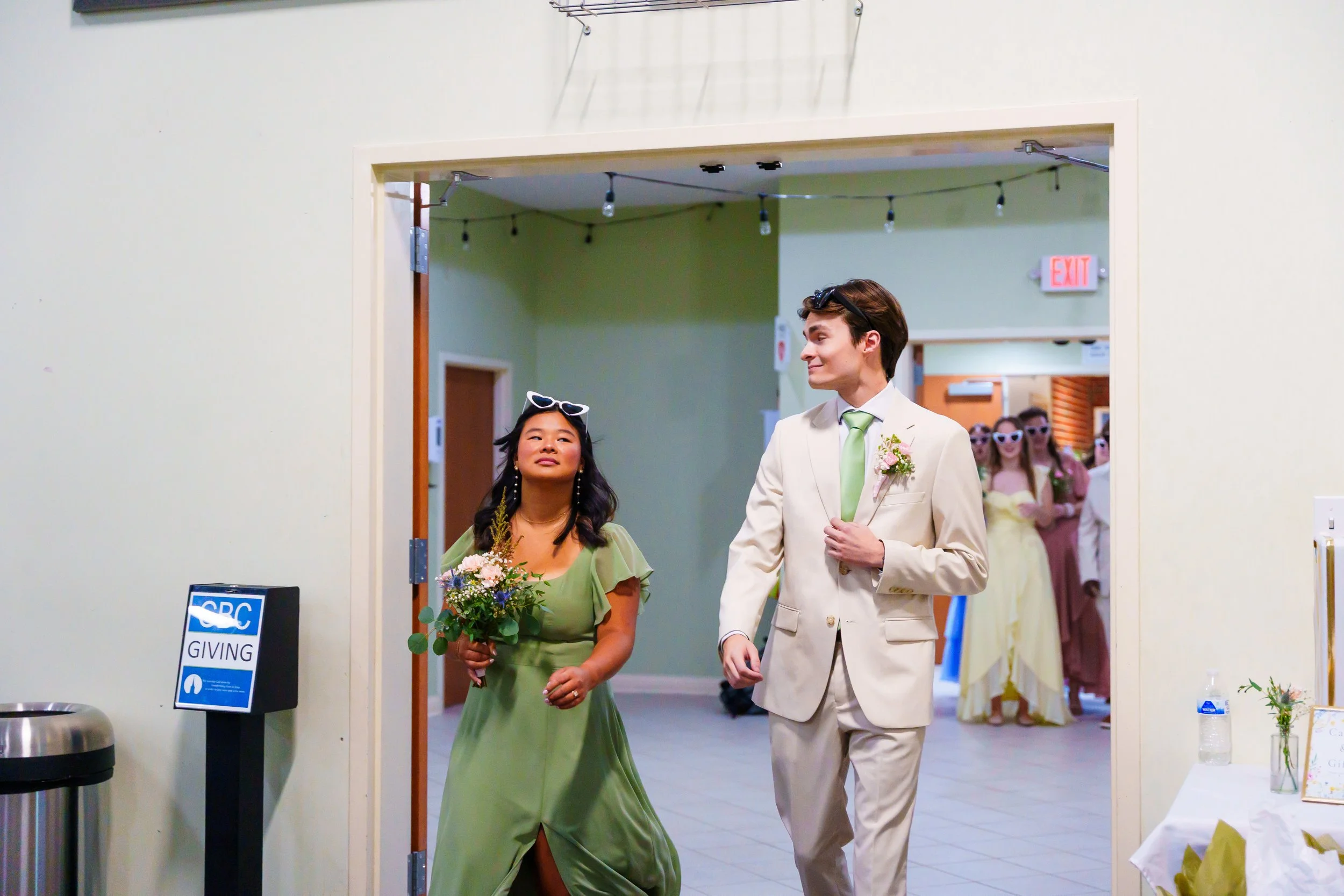 A man and woman are walking through a decorated indoor event space. The woman is wearing a green dress and holding a bouquet of flowers, and the man is in a cream suit with a light green tie, smiling at each other. In the background, there are people