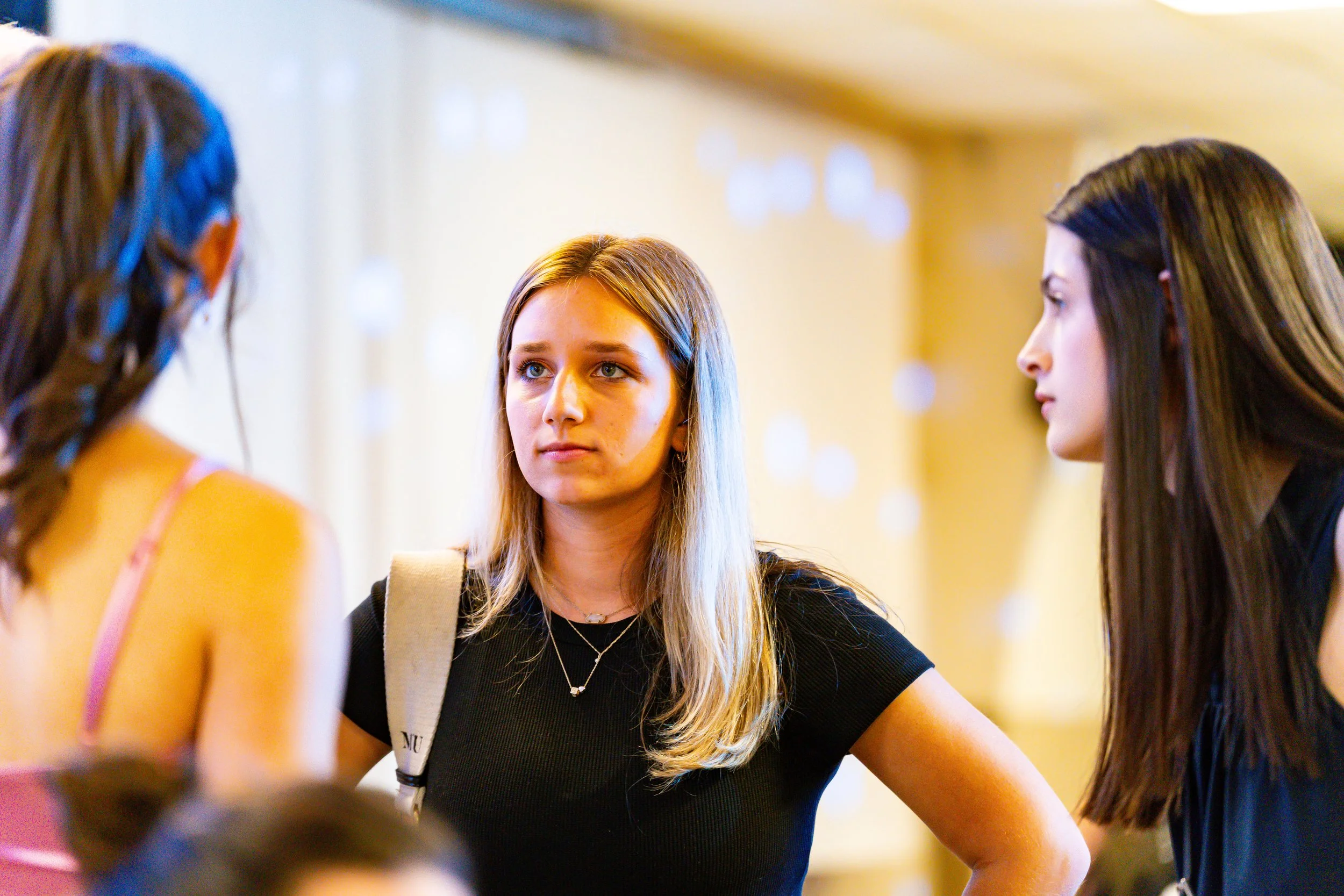 Three women are engaged in conversation at an indoor social gathering. The woman in the center has blonde hair and is wearing a black shirt, looking attentively at the woman on her left. The woman on the right has long brown hair with highlights, and