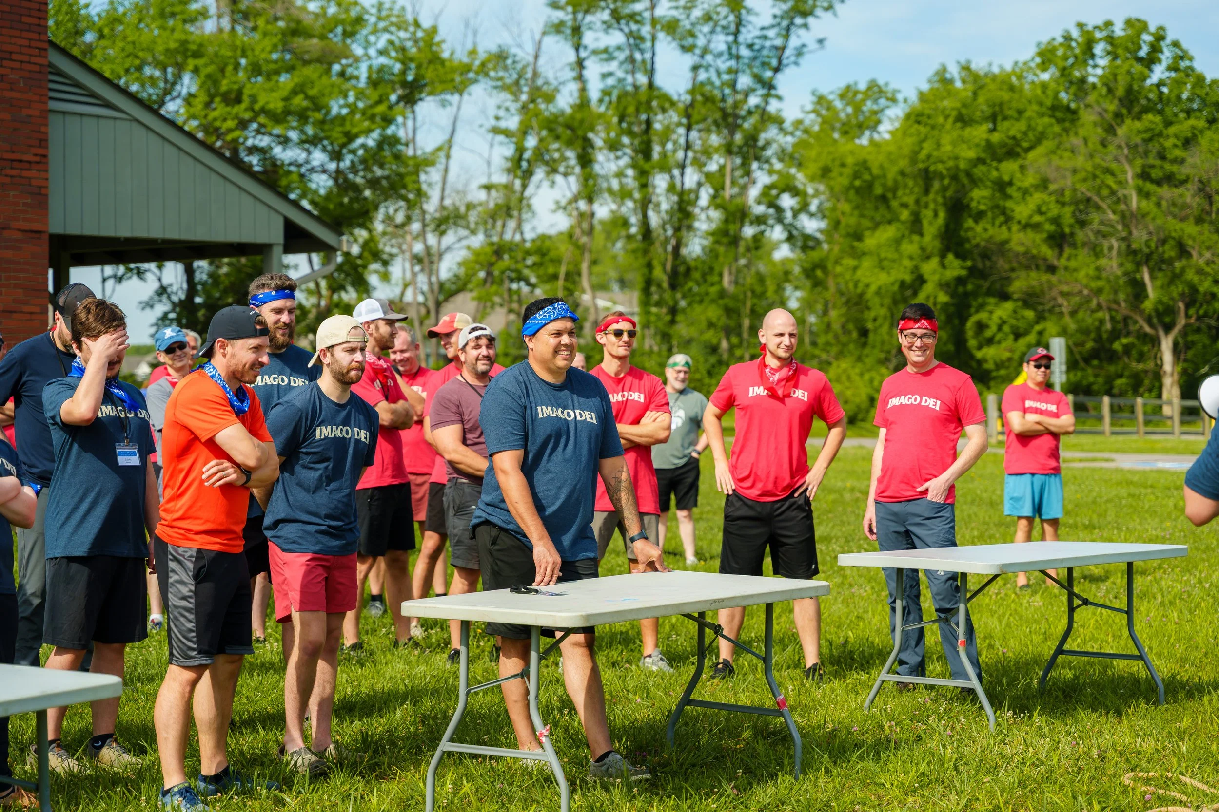 A group of people outdoors near a green area with trees, participating in a team activity or game, some wearing red and blue shirts and bandanas, and a few tables are set up on the grass.