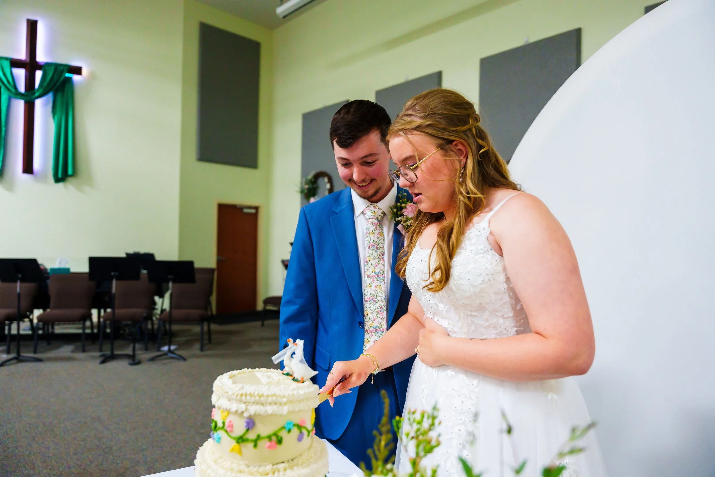 A bride and groom smiling and cutting a wedding cake inside a church or event hall with chairs and a cross on the wall in the background.