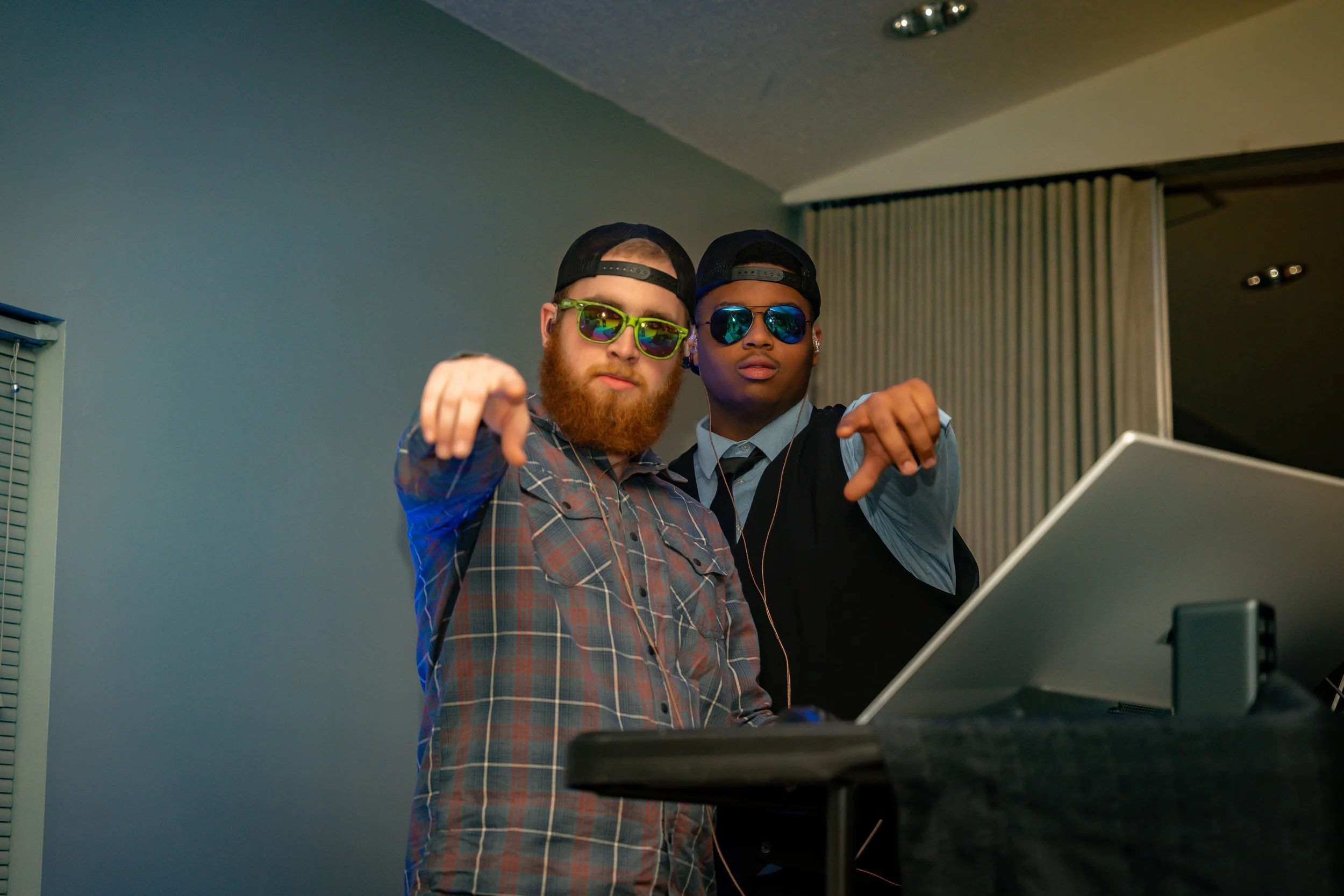 Two men wearing hats and sunglasses, one with a beard, posing indoors in front of a gray wall, pointing towards the camera, with a laptop and other electronic equipment visible in the foreground.